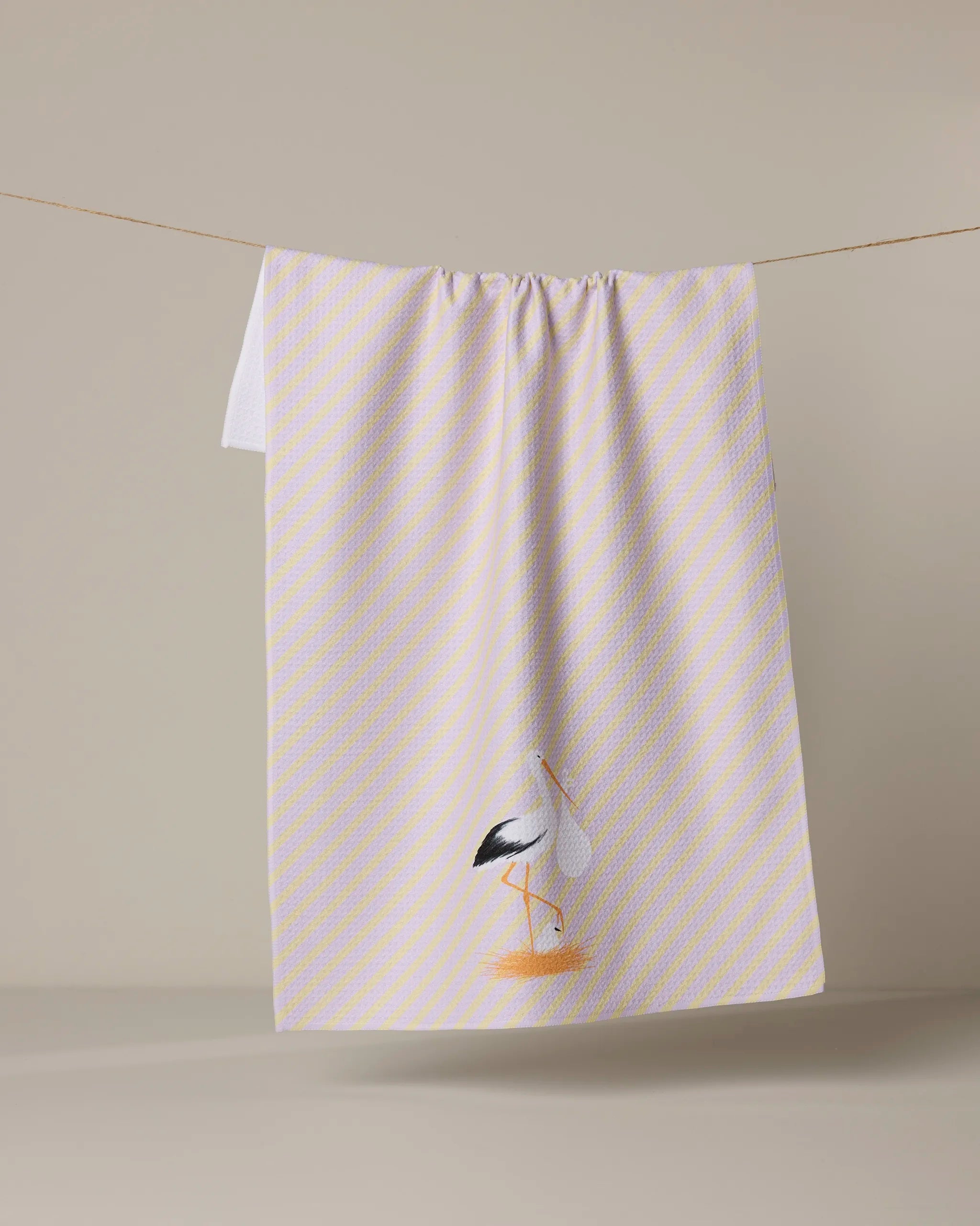 A Geometry "New Mom" dish towel in pale yellow and white with diagonal stripes hangs on a line; at the bottom, a bird illustration stands out against the plain light beige background.