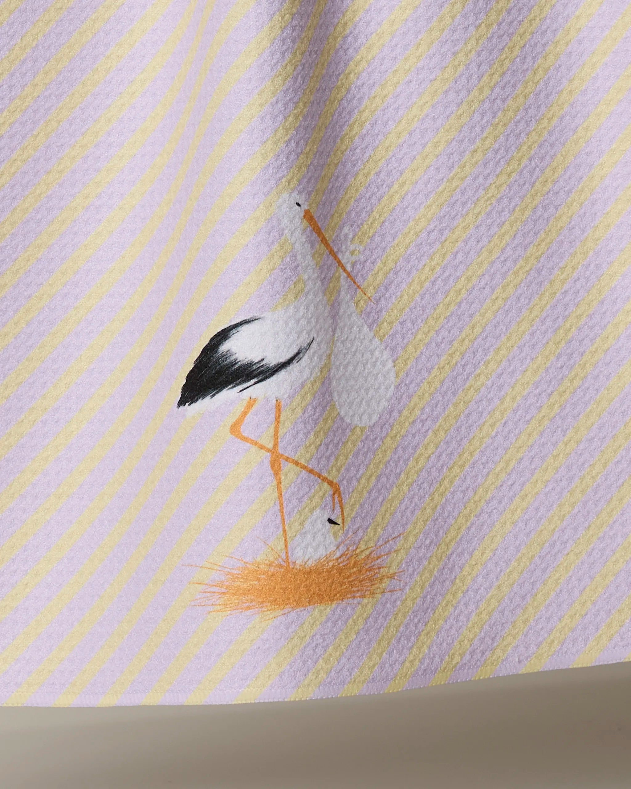 Geometry’s tea towel, “New Mom,” showcases yellow diagonal stripes on white and features an embroidered stork with orange legs and beak standing on a small orange patch.