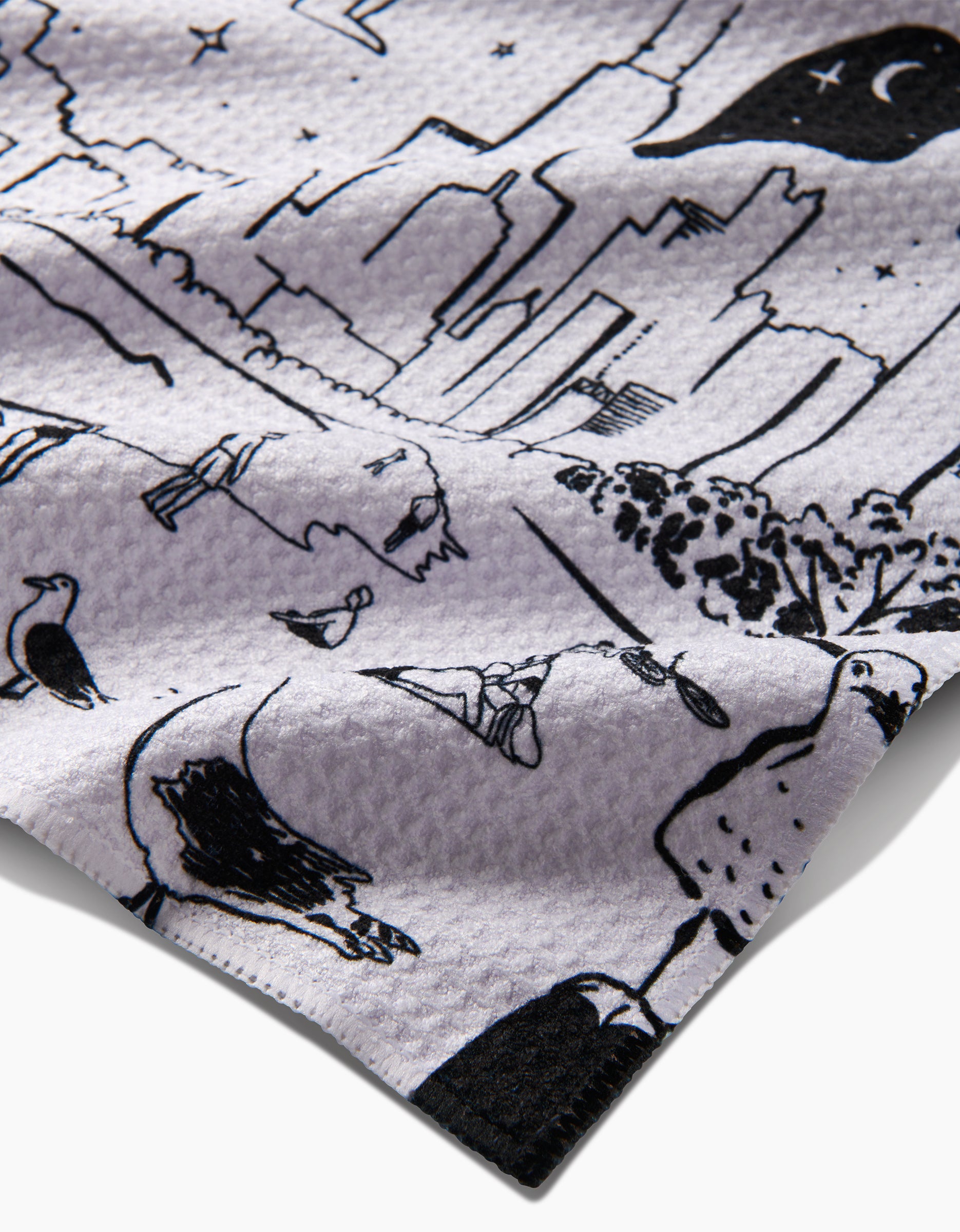 A close-up of ultra-absorbent Geoweave fabric by Elisabeth Wing, New York, featuring black city skyline, crescent moon, trees, birds, and abstract shapes on a recycled waffle-textured surface.