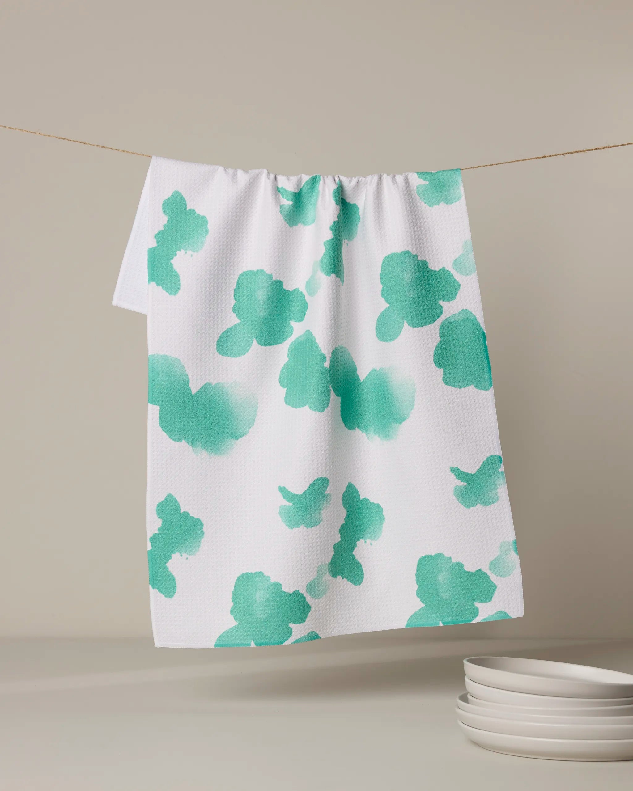 The Geometry Niamh tea towel, made from recycled materials and featuring green watercolor-like splotches on white, hangs on a string with several white plates stacked in the foreground against a neutral background.