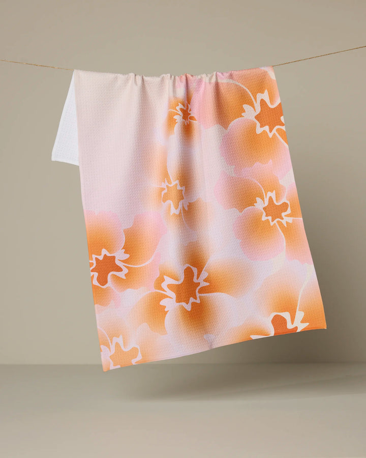 The Geometry Nicole tea towel features a pastel floral design in orange, pink, and white, and is displayed hanging against a neutral beige background.