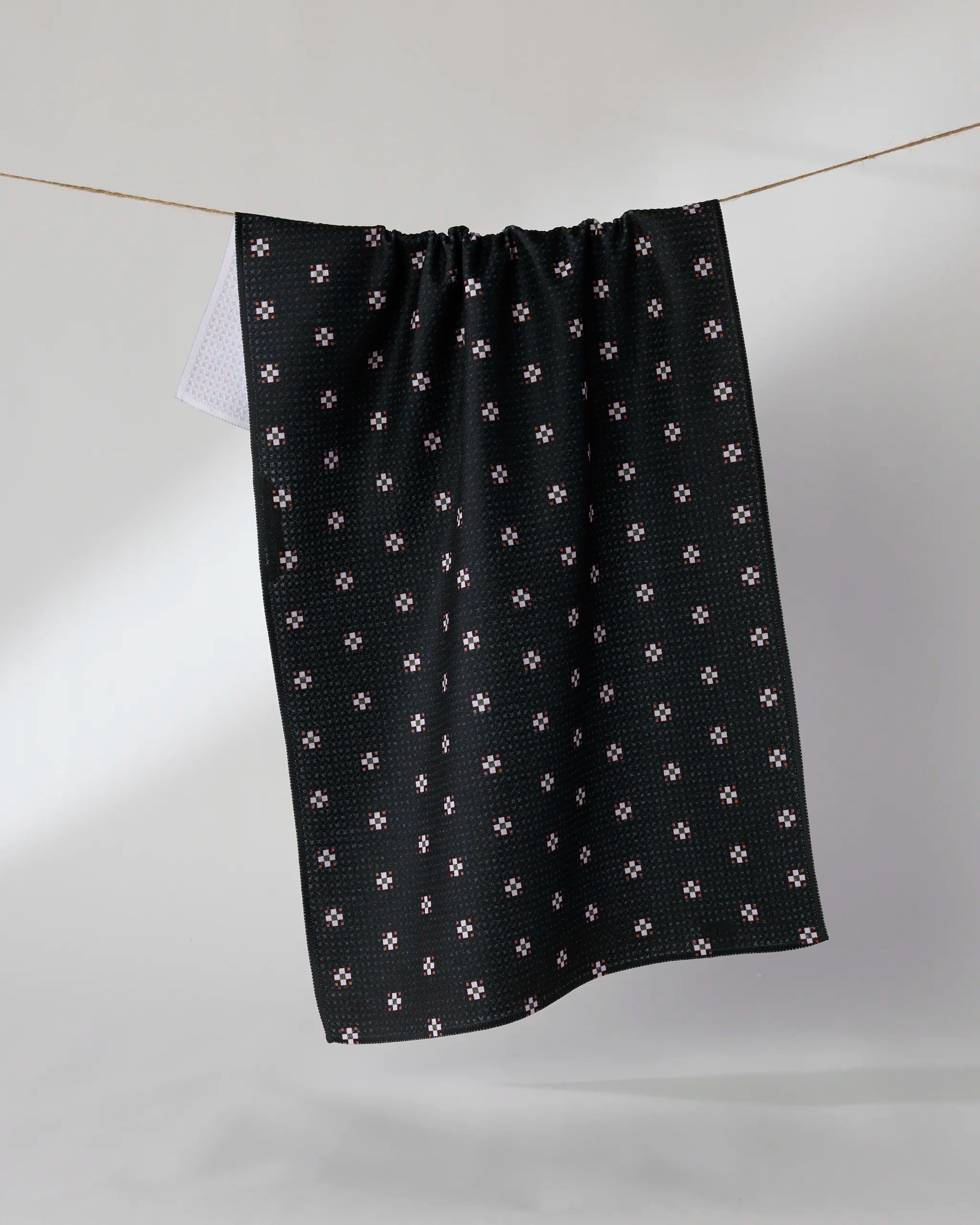 The Geometry Night Bloom tea towel, featuring a small white and pink floral pattern on black and made from recycled materials, hangs on a clothesline against a plain light background.