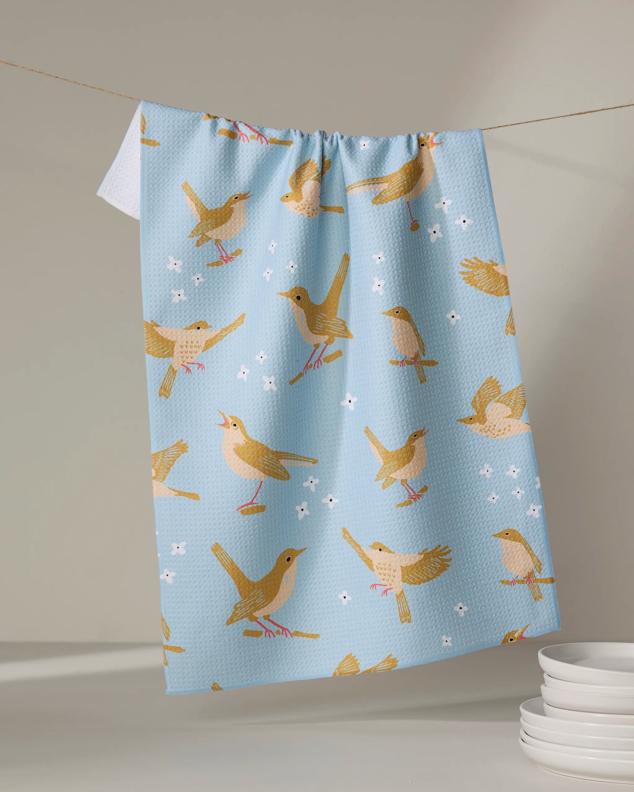 A light blue eco-friendly "Nightingales" kitchen towel by Elisabeth Wing, featuring yellow and brown birds with white flowers, hangs on a line next to a neat stack of white plates.