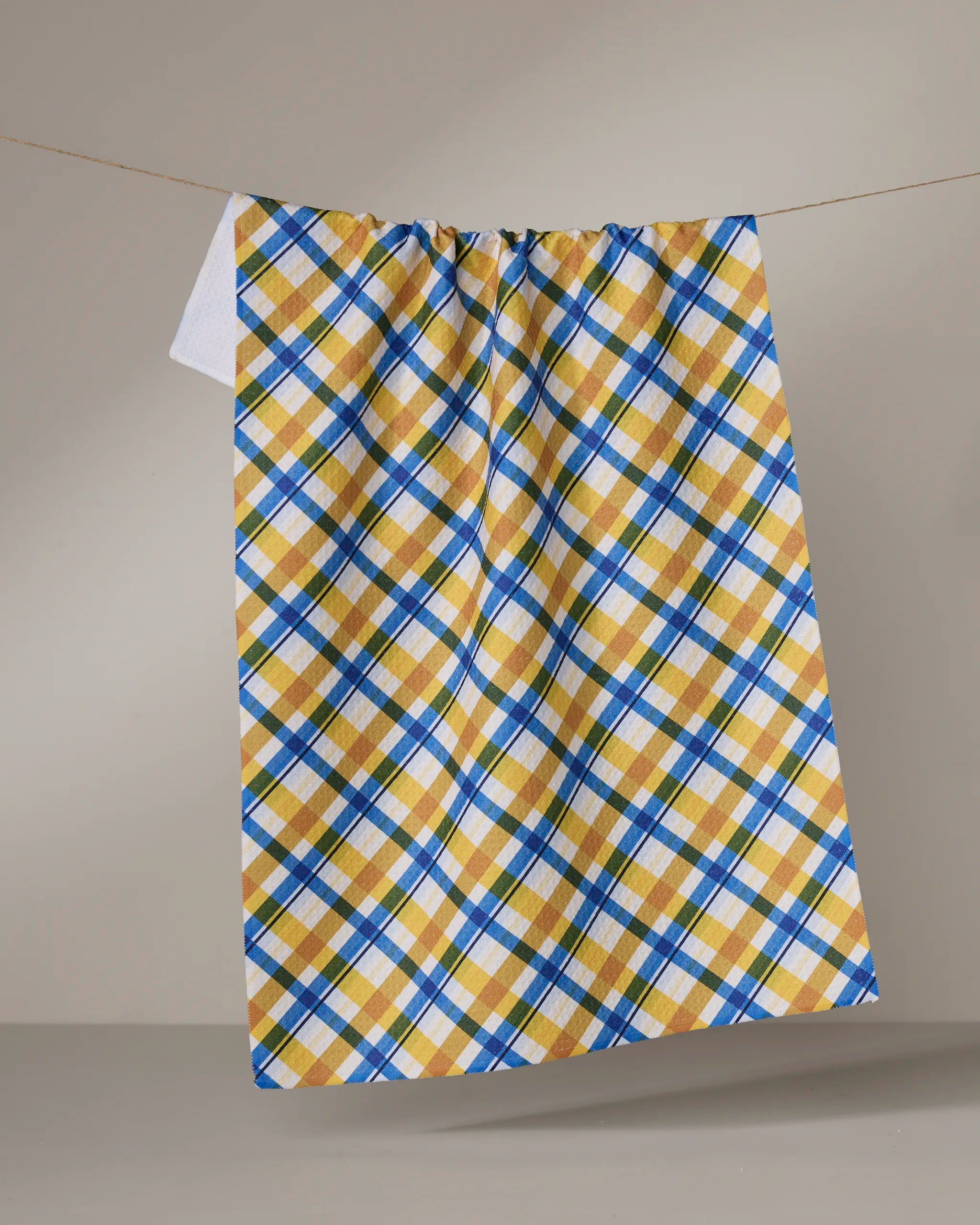 The Geometry Nochut towel, featuring a yellow, blue, and white plaid pattern, hangs on a clothesline against a neutral background. Its quick-drying design makes it both stylish and practical.