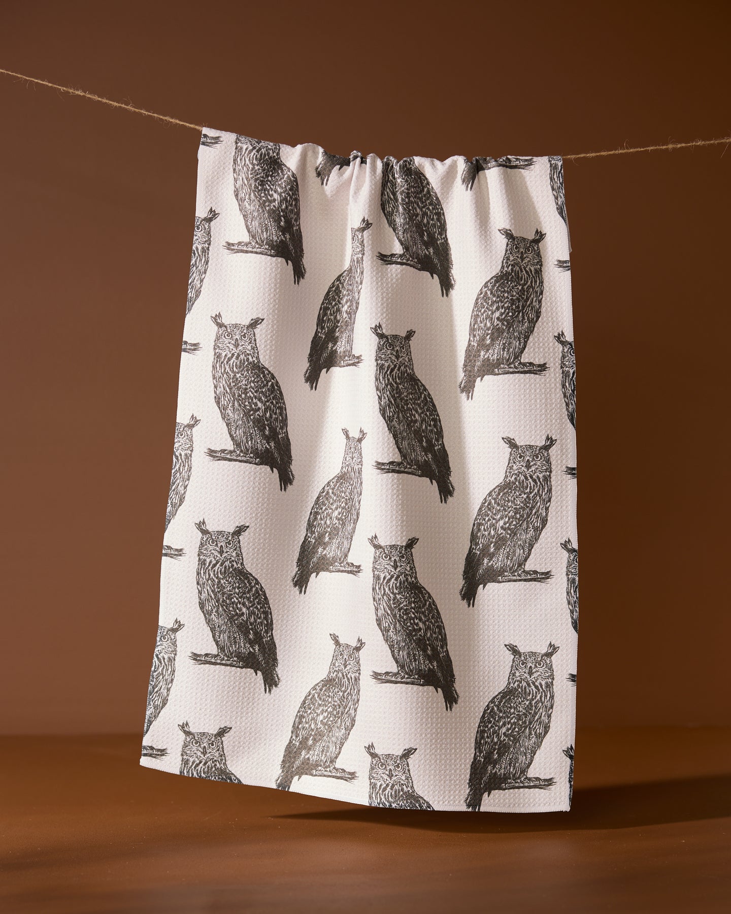 The Geometry Nocturne tea towel, featuring a repeating black owl print on white, hangs against a plain brown background.