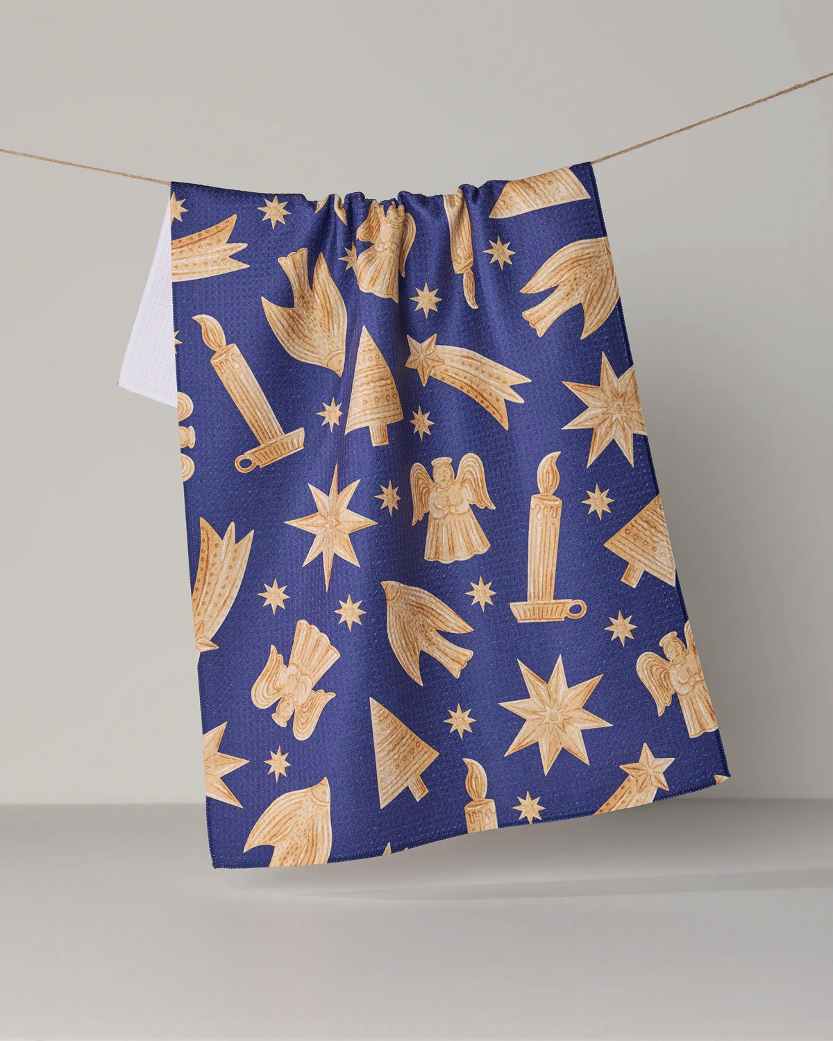 The “Noel Icons” tea towel by Geometry features golden Christmas-themed patterns—stars, candles, angels, and trees—set against a dark blue background. Made from recycled materials and ultra-absorbent for everyday use, it is pictured draped over a clothesline with a plain, light backdrop.