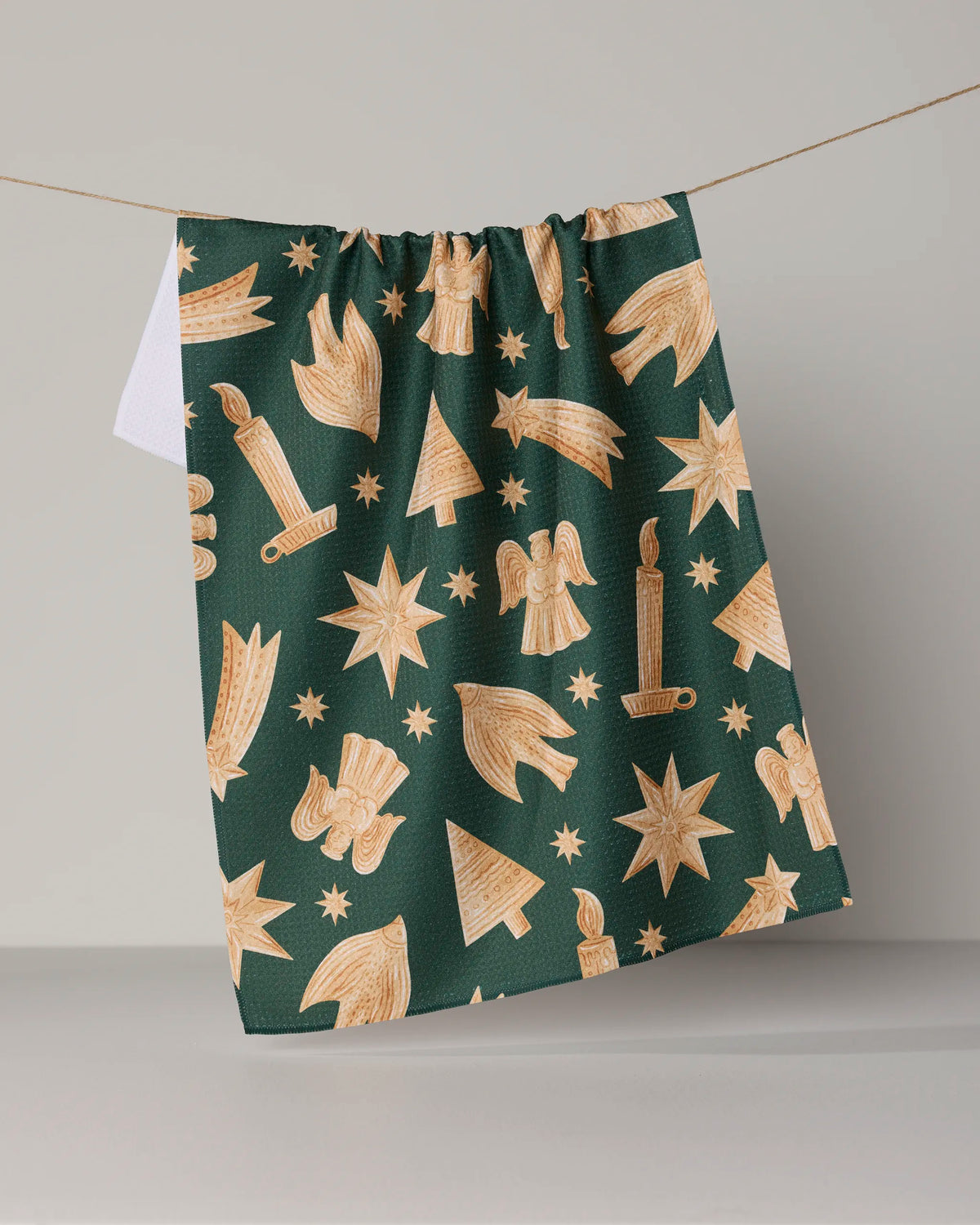 The Geometry Noel Icons kitchen tea towel hangs on a string against a plain background, showcasing a festive motif of gold stars, candles, angels, Christmas trees, and comets. Made from recycled materials, this ultra-absorbent towel combines sustainability with holiday charm for your seasonal decor.