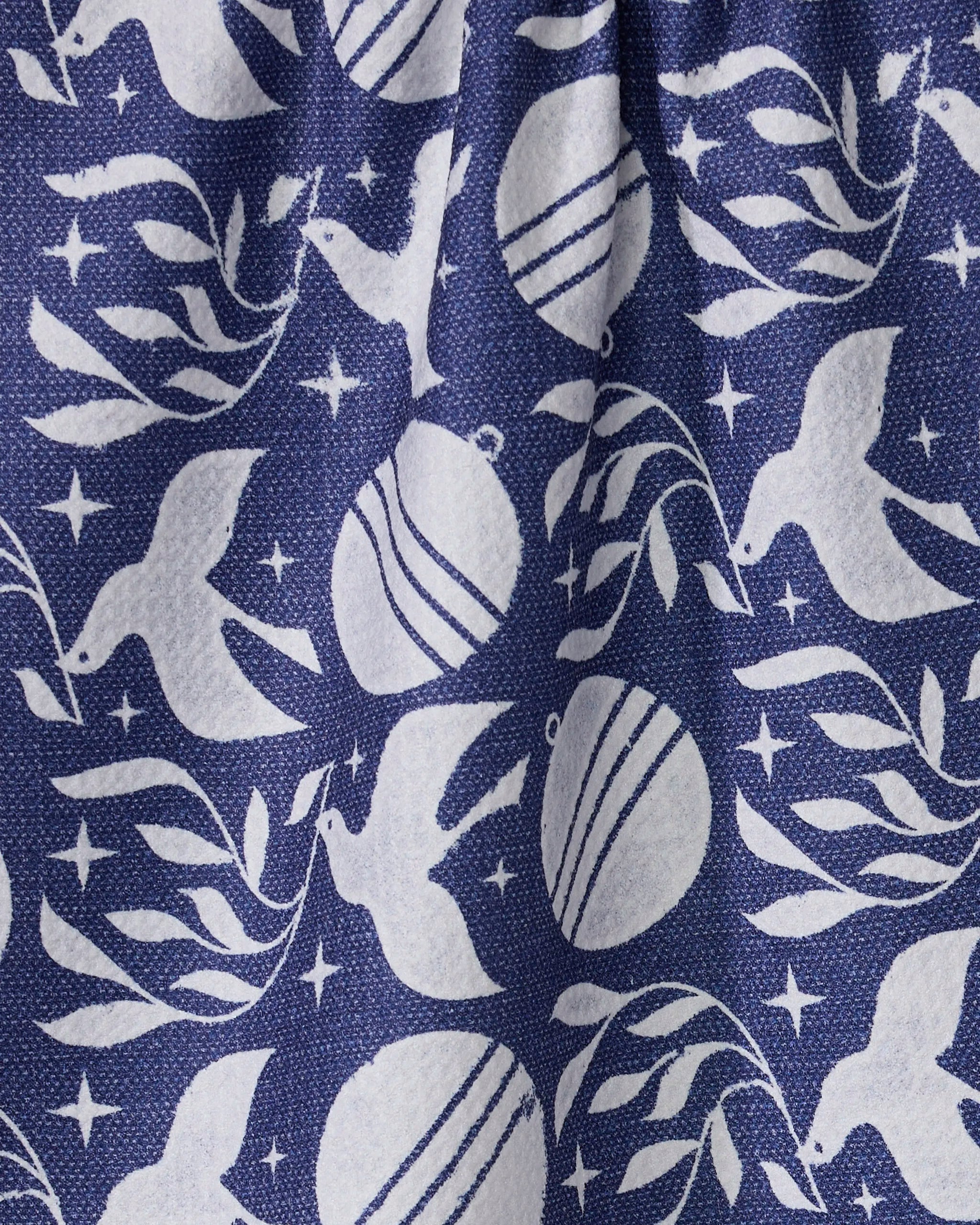 The Noel Night kitchen towel by Geometry features a repeating pattern of white doves, ornaments, and leafy branches on blue textured fabric made from recycled materials, with gentle folds visible.