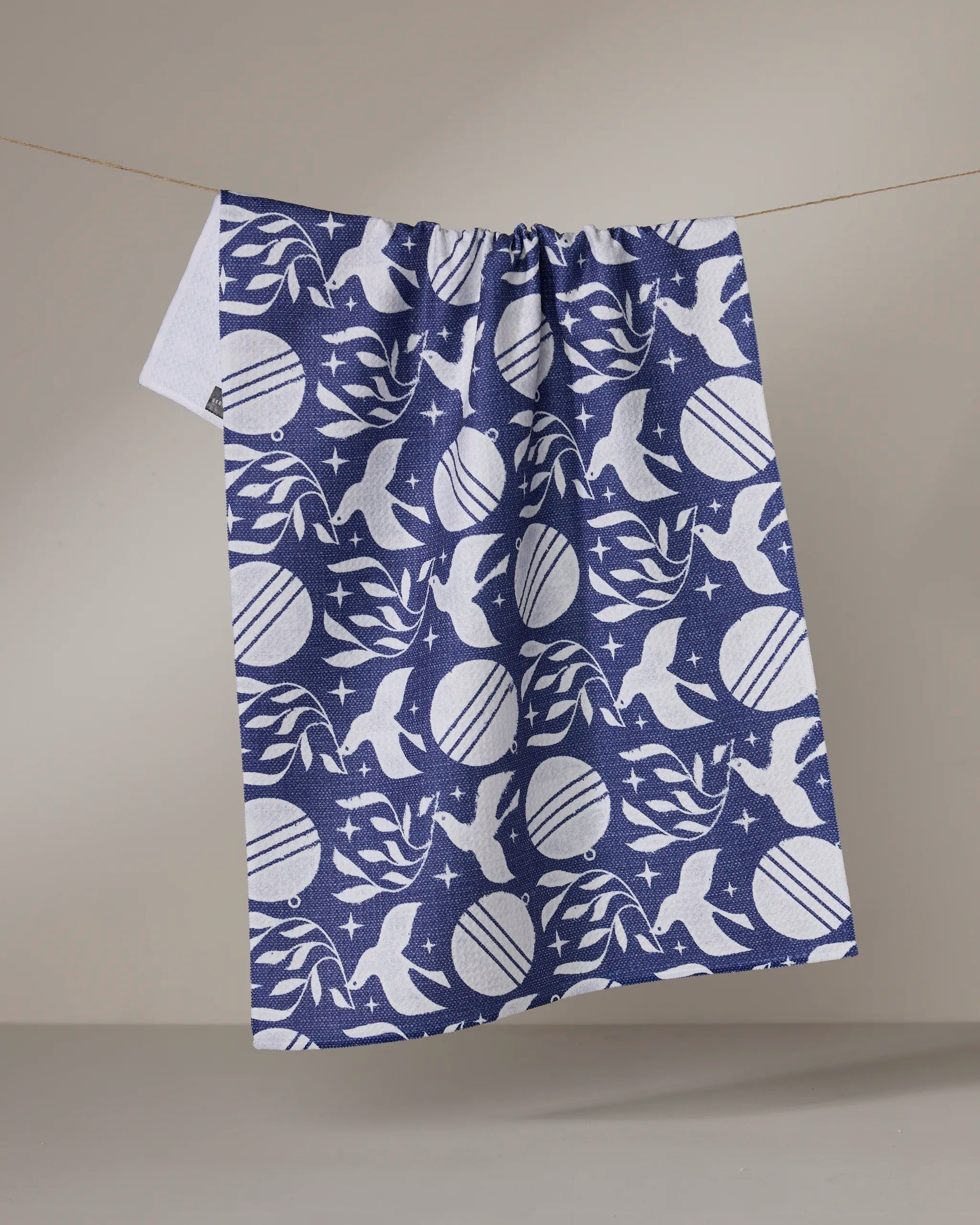 The Geometry Noel Night tea towel hangs from a string against a plain gray background, showcasing blue and white birds, leaves, stars, and round shapes.