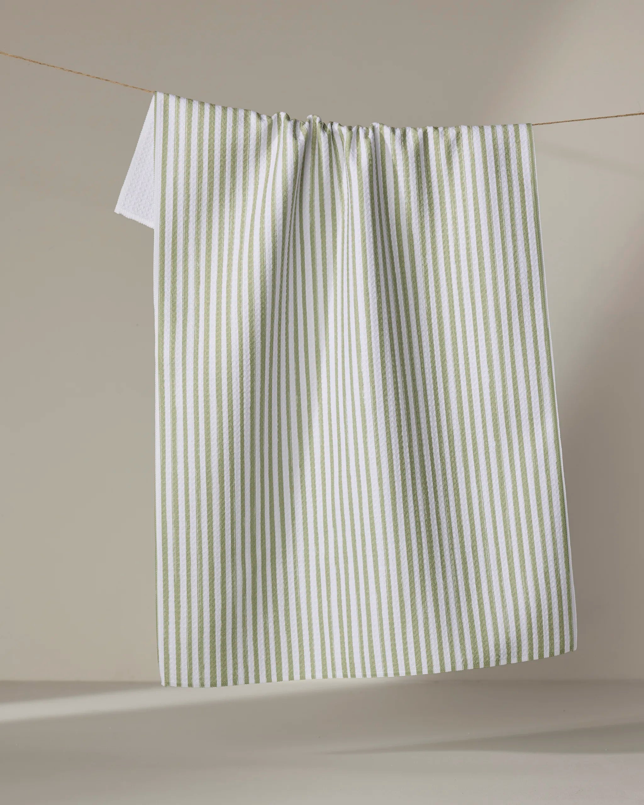 The Noel kitchen towel by Kailee Wright, made from recycled materials, features vertical light green stripes on white and hangs neatly on a clothesline against a plain beige background.
