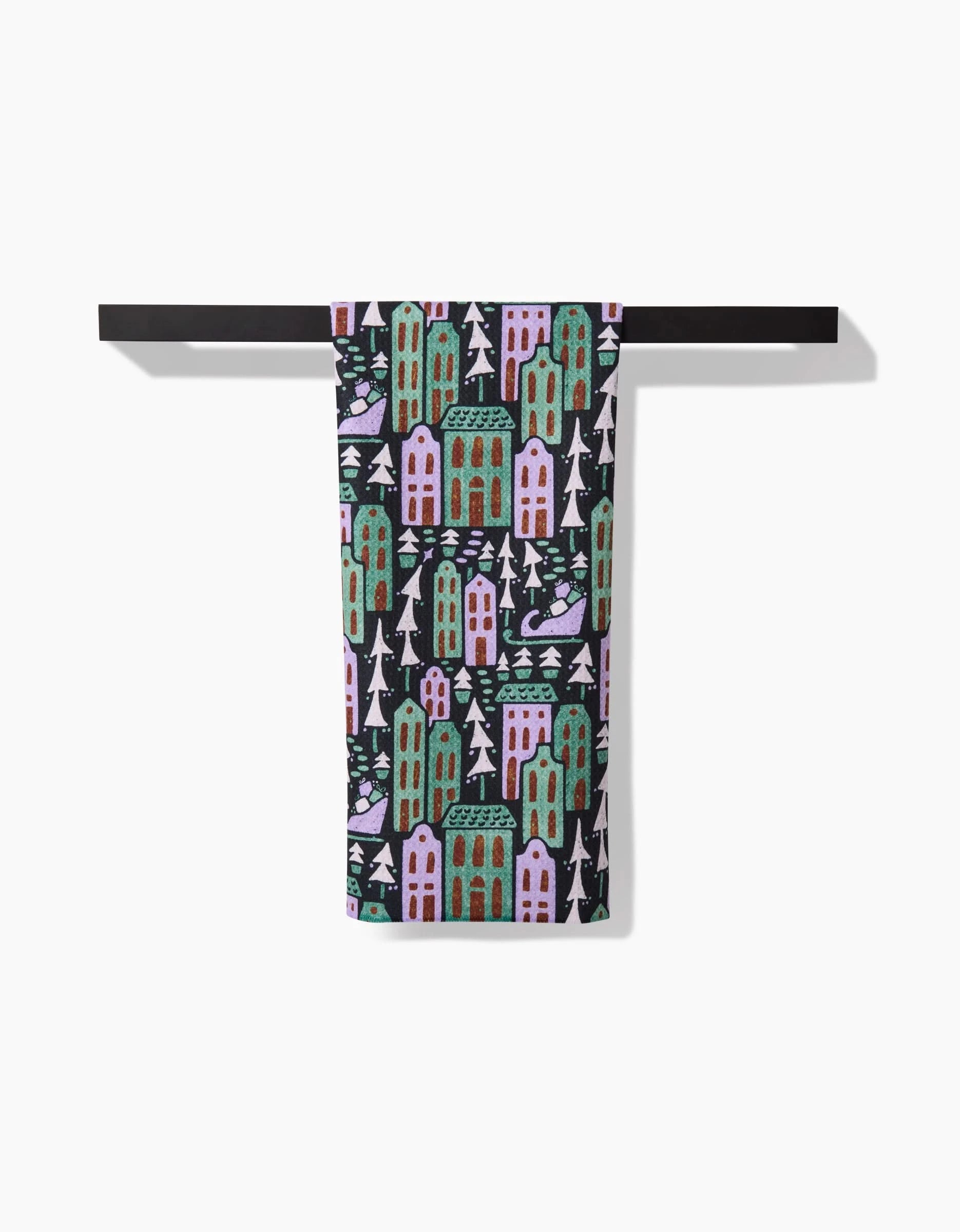 The Noelle tea towel by Amy MacCready hangs on a black bar, showcasing pink and green buildings, white trees, and blue accents on dark fabric. Crafted from ultra-absorbent recycled materials for style and sustainability.