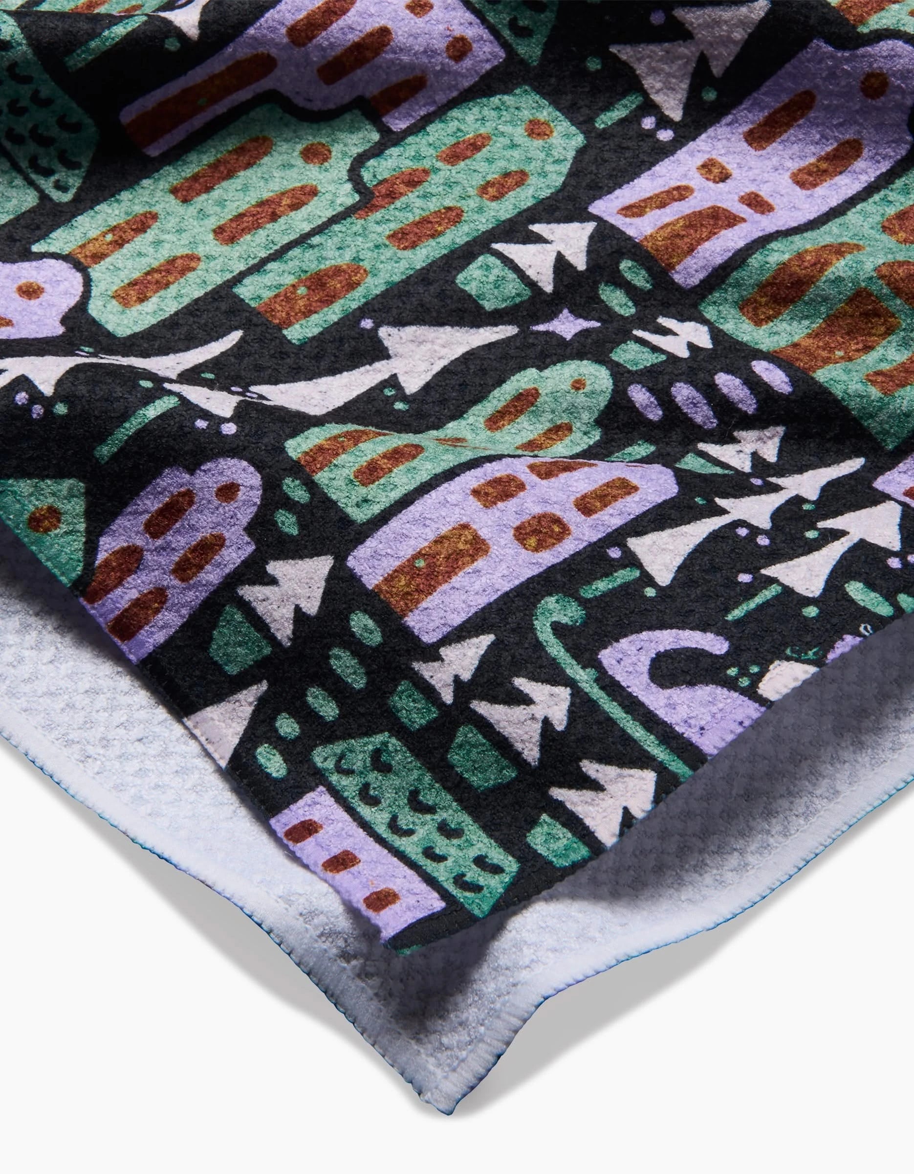 A close-up of the Noelle fleece blanket by Amy MacCready, made from recycled materials, displays a colorful abstract pattern of houses, trees, and shapes in green, purple, brown, and white on black; edges and underside are shown.