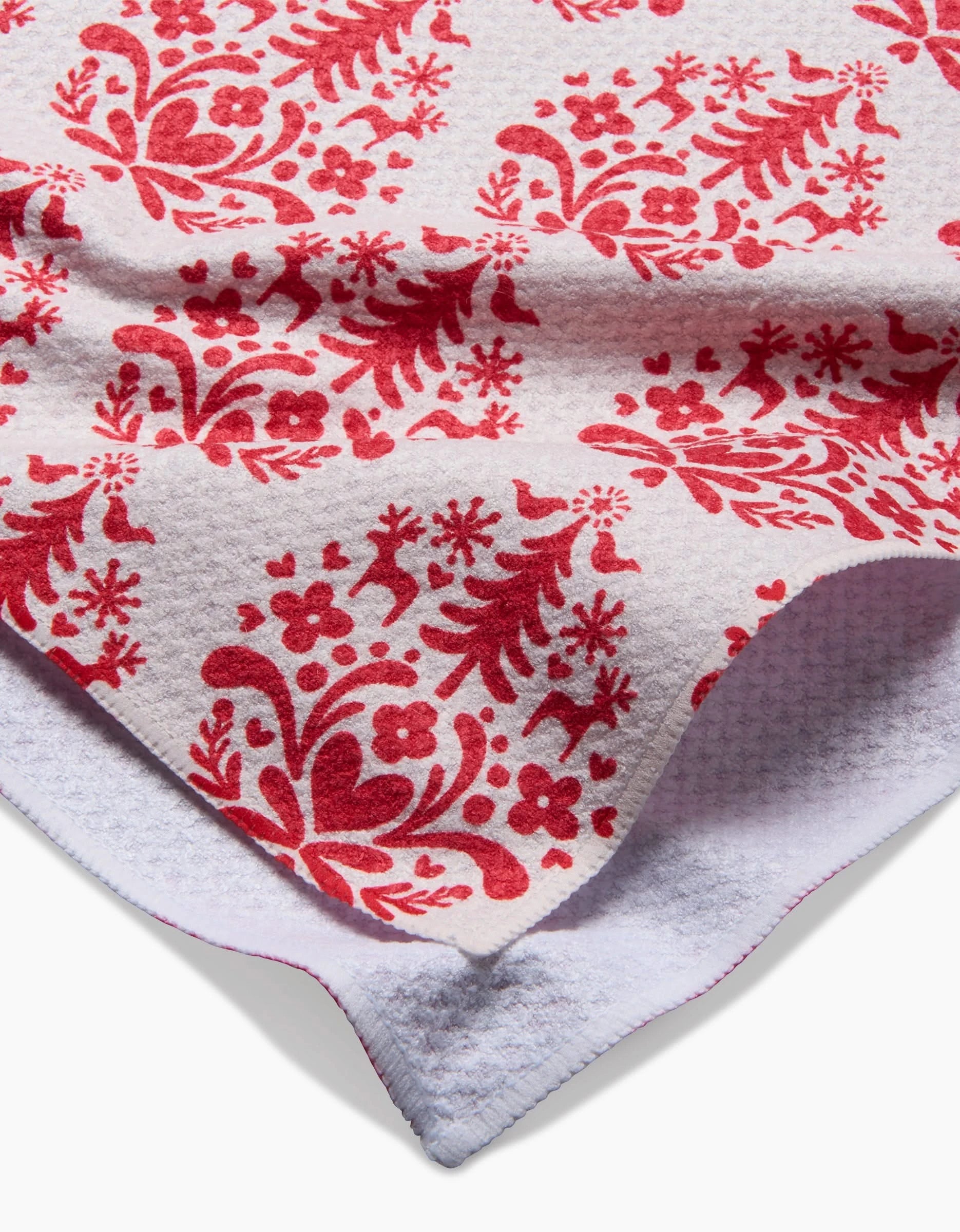 A close-up of Kayla Ann’s Nordic Holiday Stamp tea towel shows its ultra-absorbent red floral and animal pattern on white, with a folded corner revealing a textured underside and red stitched edge.