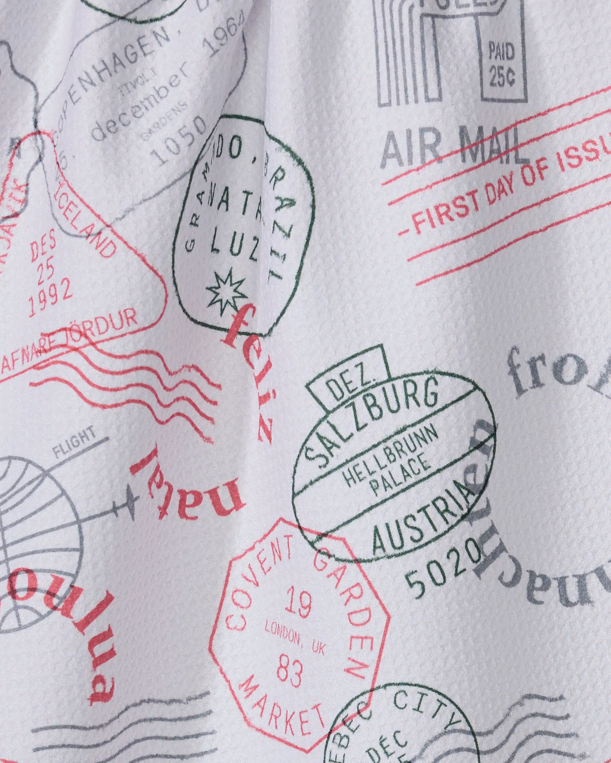 The Geometry Nordic Mail tea towel features passport stamp and postmark prints in red, black, and green with locations like Salzburg, Covent Garden Market, Iceland, and Brazil. Made from recycled materials with a textured surface.