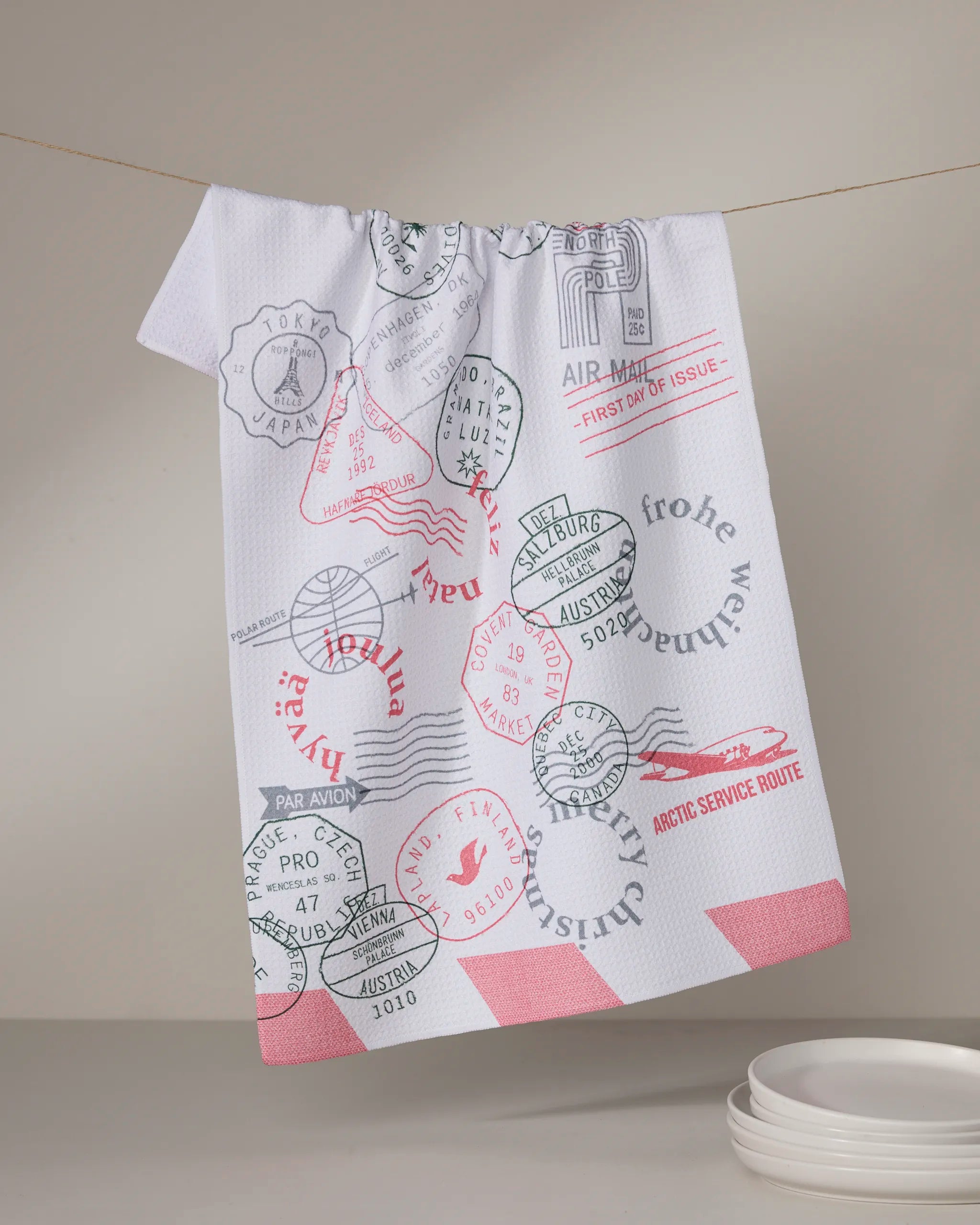 The Geometry Nordic Mail tea towel, made from recycled materials, features colorful passport stamp designs and hangs above white plates against a plain light background.