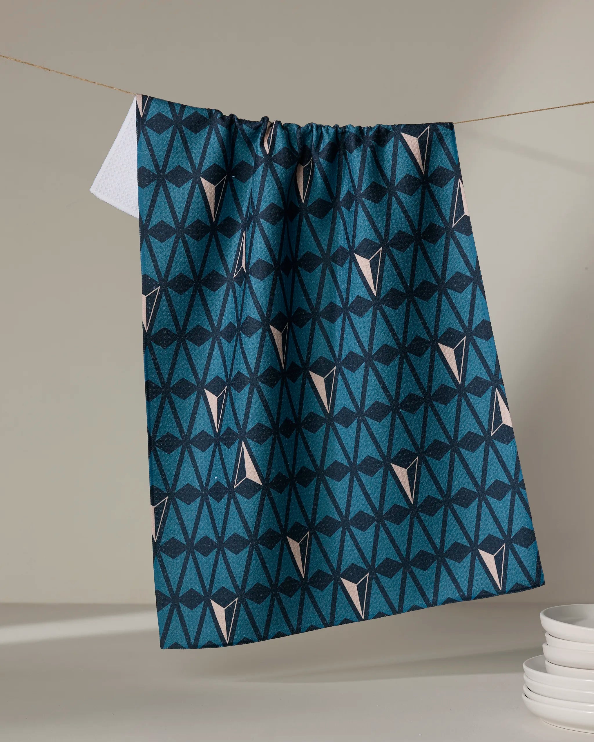 The Northbound tea towel by Twig + Spark, featuring a blue and black geometric pattern and made from recycled materials, hangs on a clothesline beside stacked white plates on a neutral background.