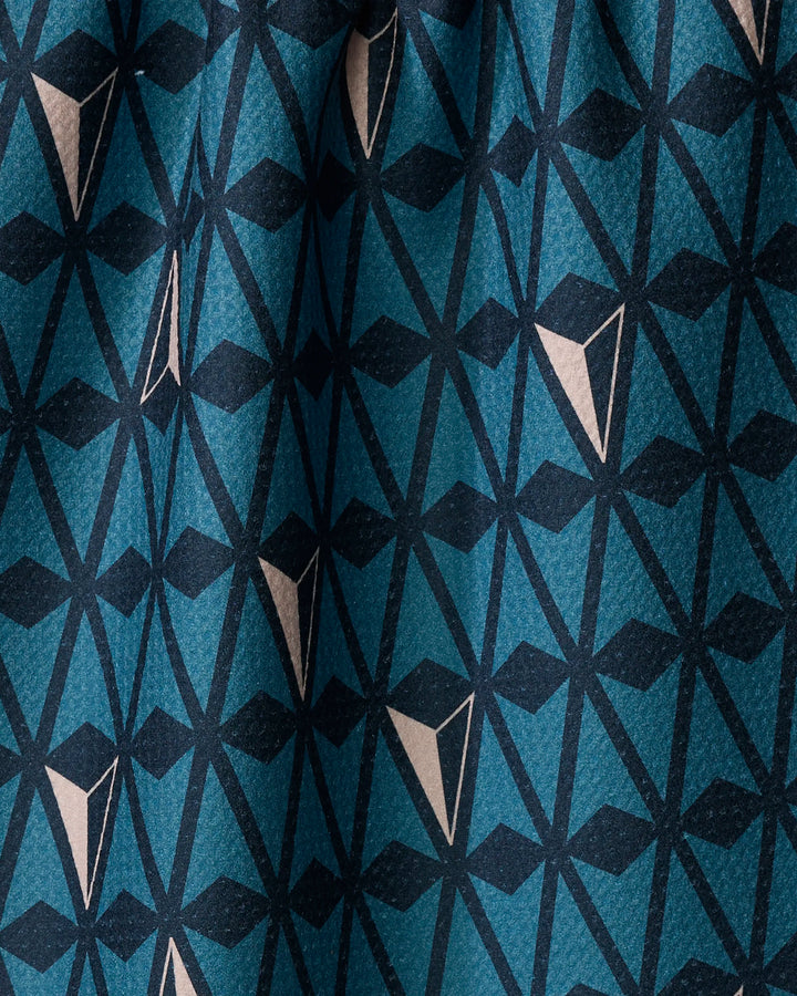 A close-up of Twig + Spark's Northbound tea towel shows its quick-drying fabric with a dynamic geometric pattern in teal, navy, and beige triangles and lines. The repeating design appears slightly folded, perfect for modern kitchens.