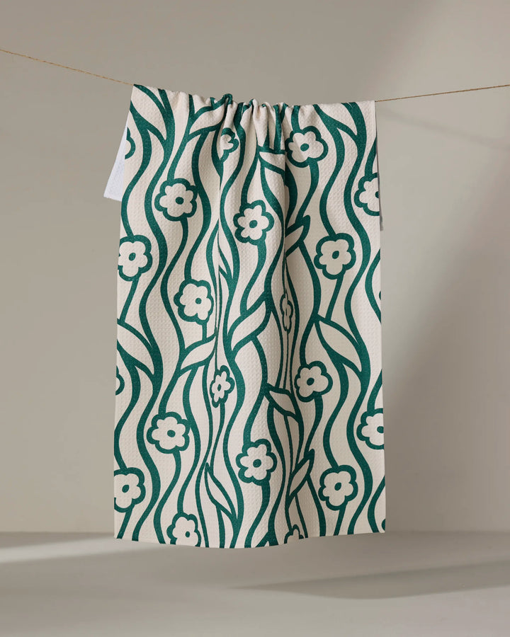 The Gerdadzy Nostalgic Flowers tea towel, cream-colored with a green floral and vine design and made from recycled materials, hangs on a clothesline against a plain light background.