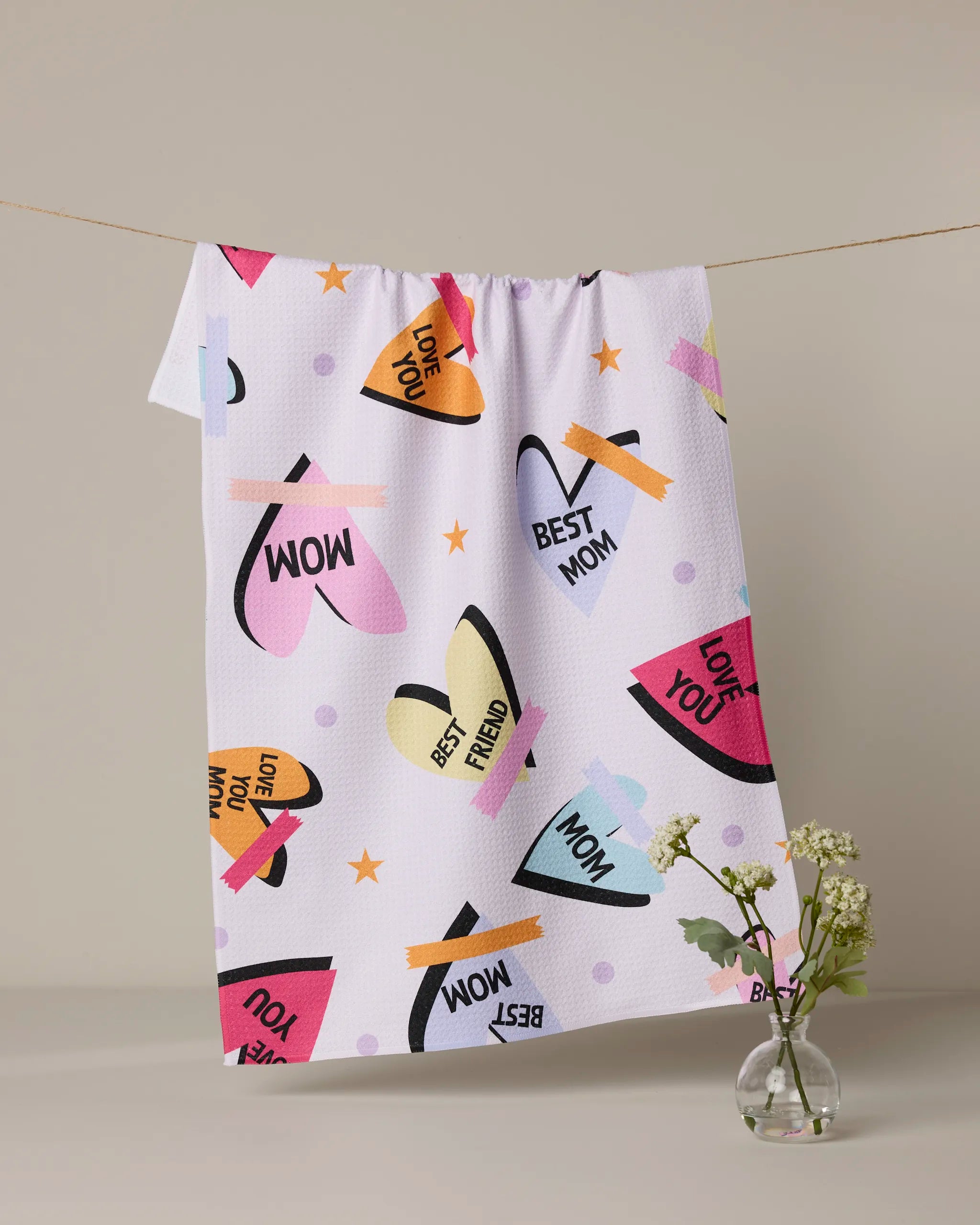 The Geometry “Notes to Mom” dish towel, decorated with heart shapes and cheerful messages, hangs on a line. Beneath it, a clear vase of white flowers sits against a neutral backdrop.