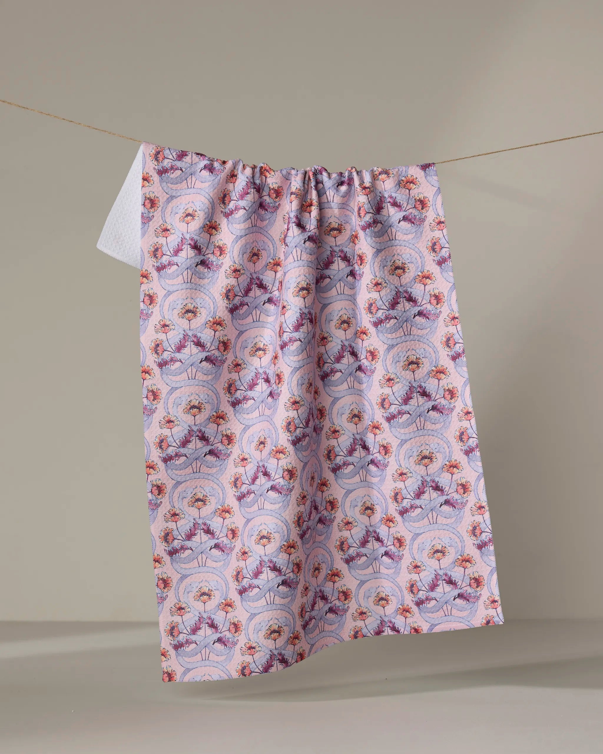 The Nouveau Snake kitchen towel by Maayan Alper-Swan, featuring eco-friendly fabric and vibrant pink, purple, and red floral and geometric patterns, hangs on a line against a neutral beige background.