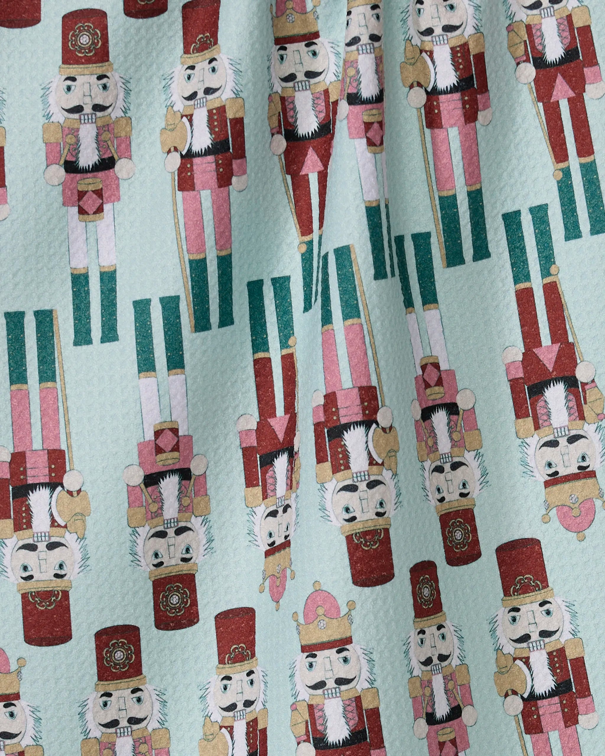 The Nutcracker March kitchen towel by Geometry features illustrated nutcrackers in red, pink, and gold on a light blue background. Made with recycled materials, the playful pattern includes both upright and upside-down nutcrackers.