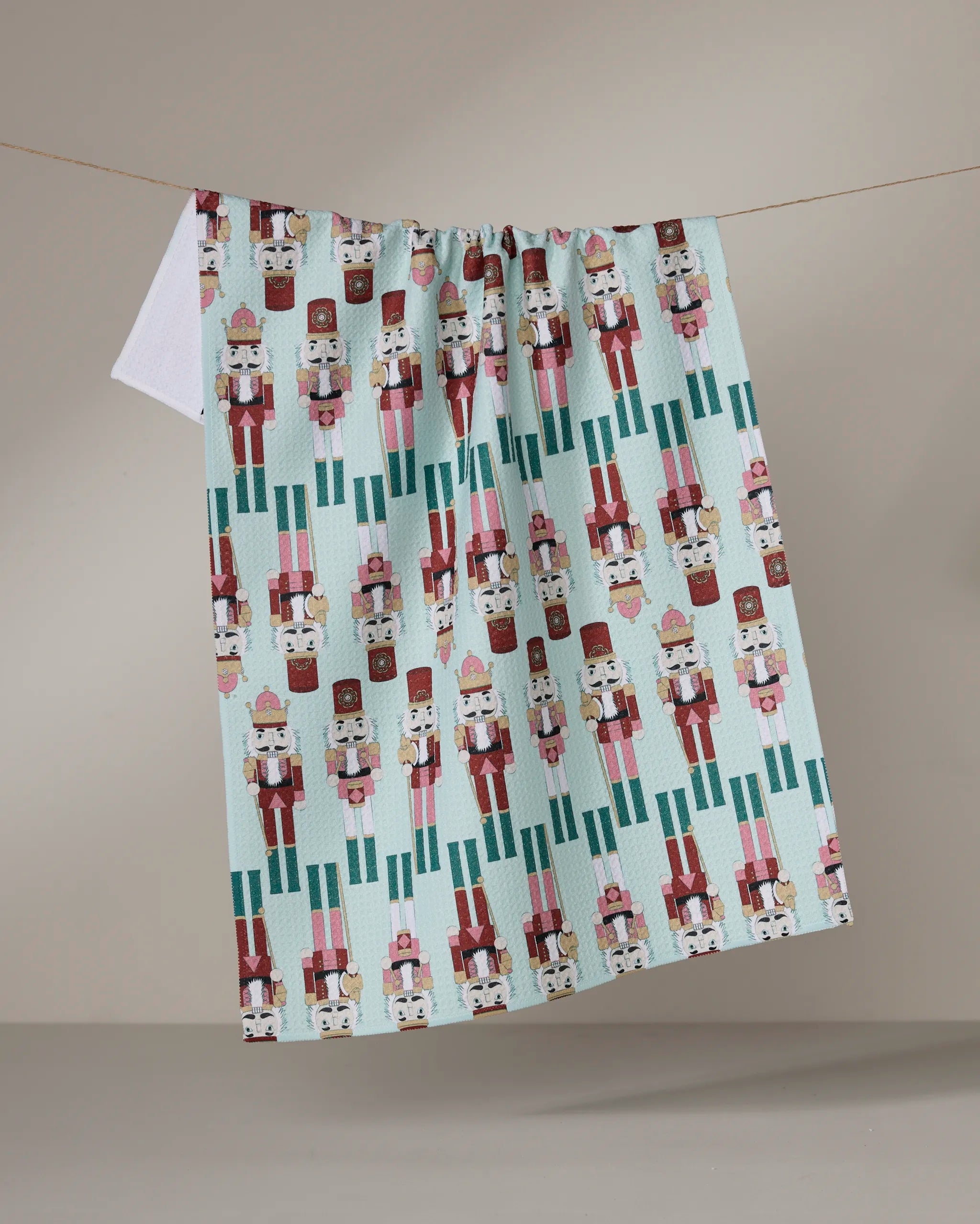 The Geometry "Nutcracker March" tea towel, with a colorful nutcracker pattern in red, pink, teal, and white, hangs on a line against a plain beige background.