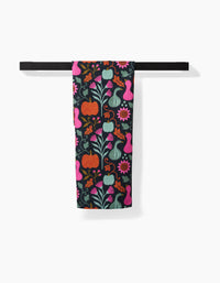 The Oakley kitchen towel by Amy MacCready, made from recycled materials and featuring pumpkins, flowers, leaves, and gourds in vibrant pink, orange, green, and blue, hangs on a black towel bar against a white background.