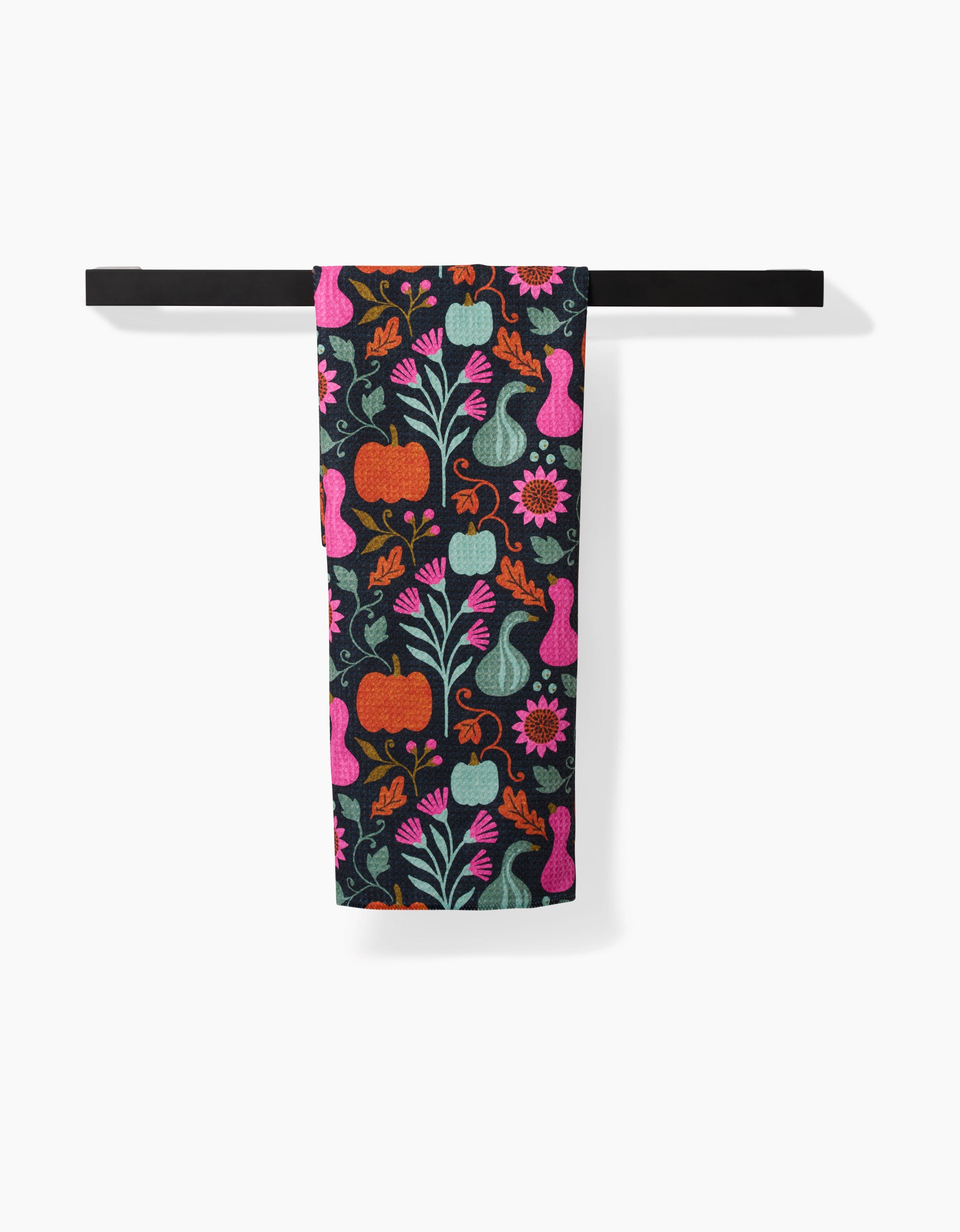 The Oakley kitchen towel by Amy MacCready, made from recycled materials and featuring pumpkins, flowers, leaves, and gourds in vibrant pink, orange, green, and blue, hangs on a black towel bar against a white background.