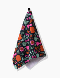 The Oakley hanging kitchen towel by Amy MacCready features a vibrant floral and fruit print in orange, pink, and green on black, crafted from recycled materials for eco-friendly style. Displayed against a white surface.