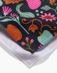 A close-up of two overlapping Amy MacCready Oakley kitchen towels: one shows a vibrant floral and fruit pattern on dark fabric, the other a plain white textured underside. Both are crafted from recycled materials for eco-friendly appeal.