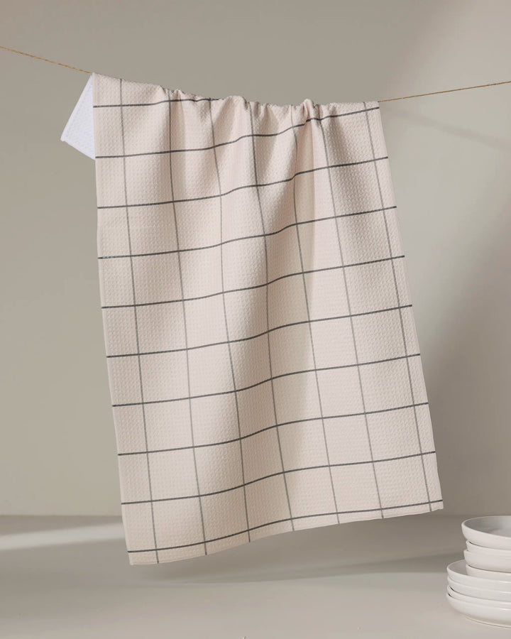 The Oat Grid tea towel by Kelsi Fullmer, featuring a beige base with a simple black grid design, hangs on a string with its white corner folded over. Stacked white plates sit in the lower right against a neutral backdrop.