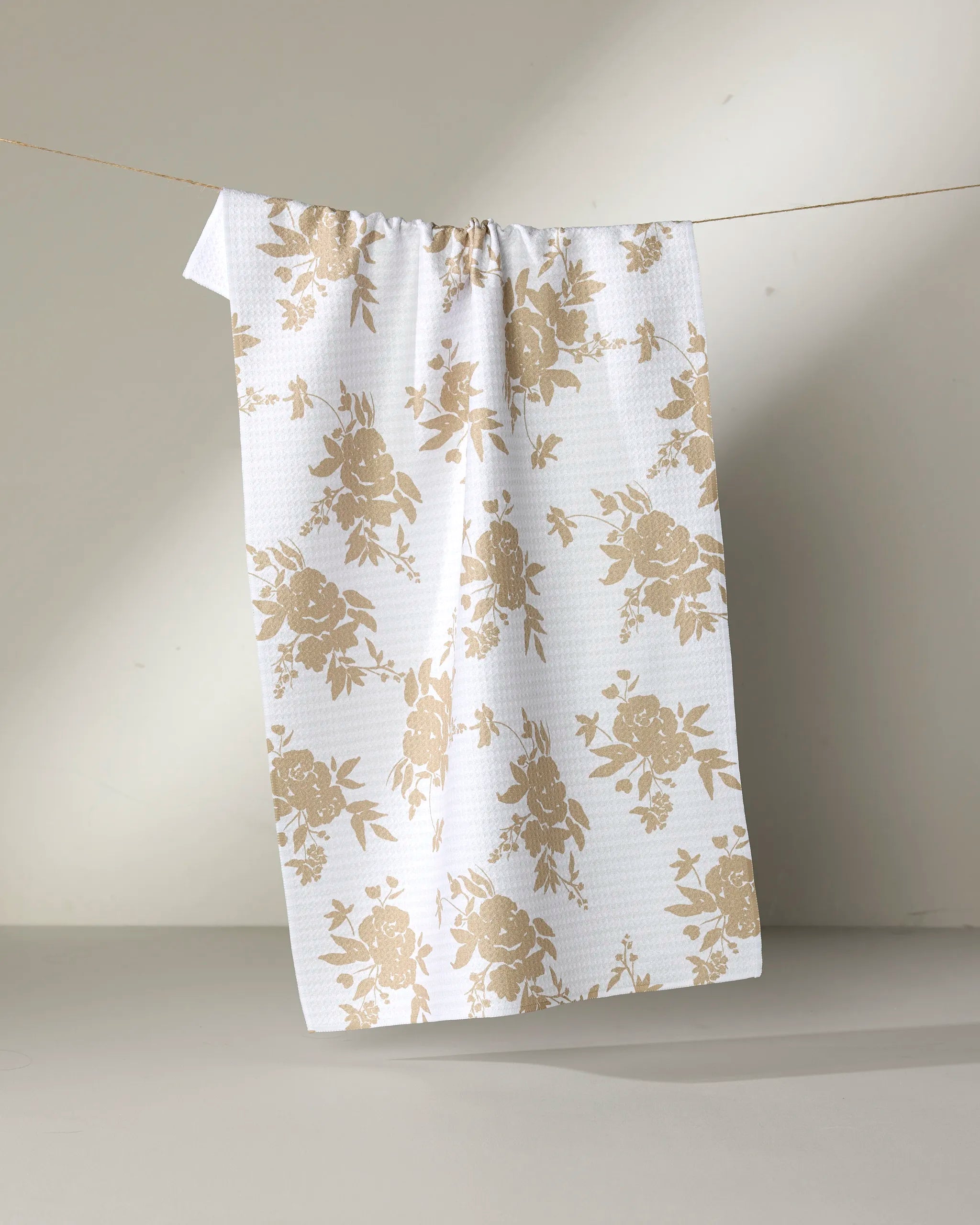 The Aubrey Fairchild “Oatmeal Floral” tea towel, featuring a beige floral pattern, hangs from a thin string against a plain, light-colored background.