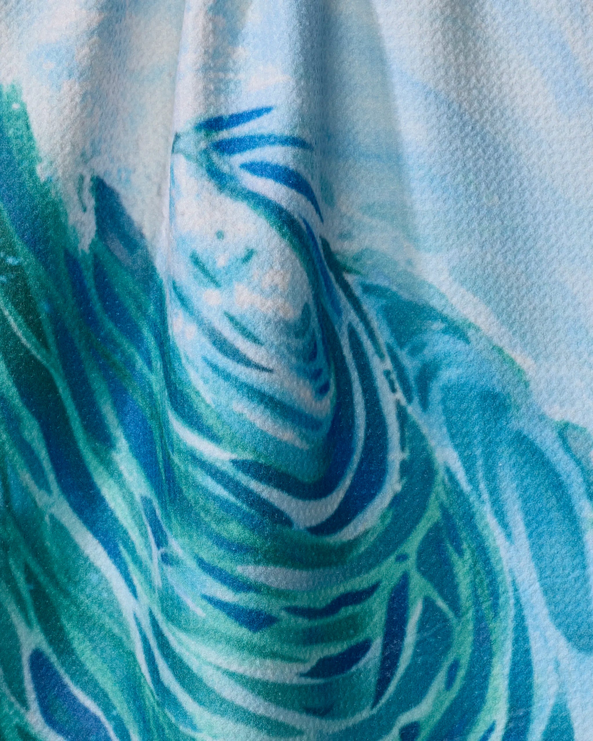 Close-up of Trey Jolley’s Ocean Wave tea towel, crafted from recycled materials. The ultra-absorbent fabric displays blue, green, and white abstract swirls inspired by ocean waves, with its textured folds clearly visible.