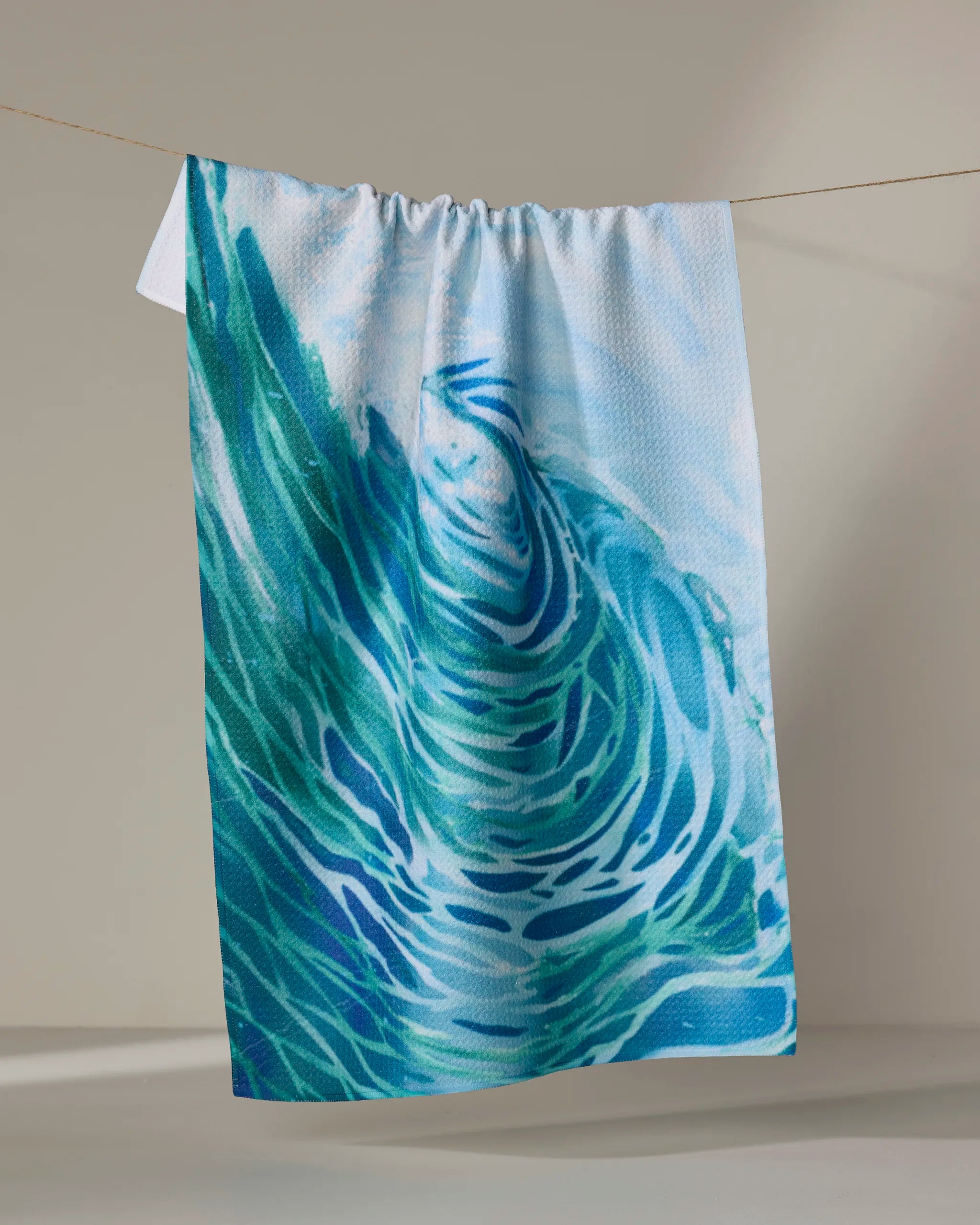 The Trey Jolley Ocean Wave tea towel, crafted from recycled materials and featuring a blue and turquoise abstract water pattern, hangs on a clothesline against a neutral background.