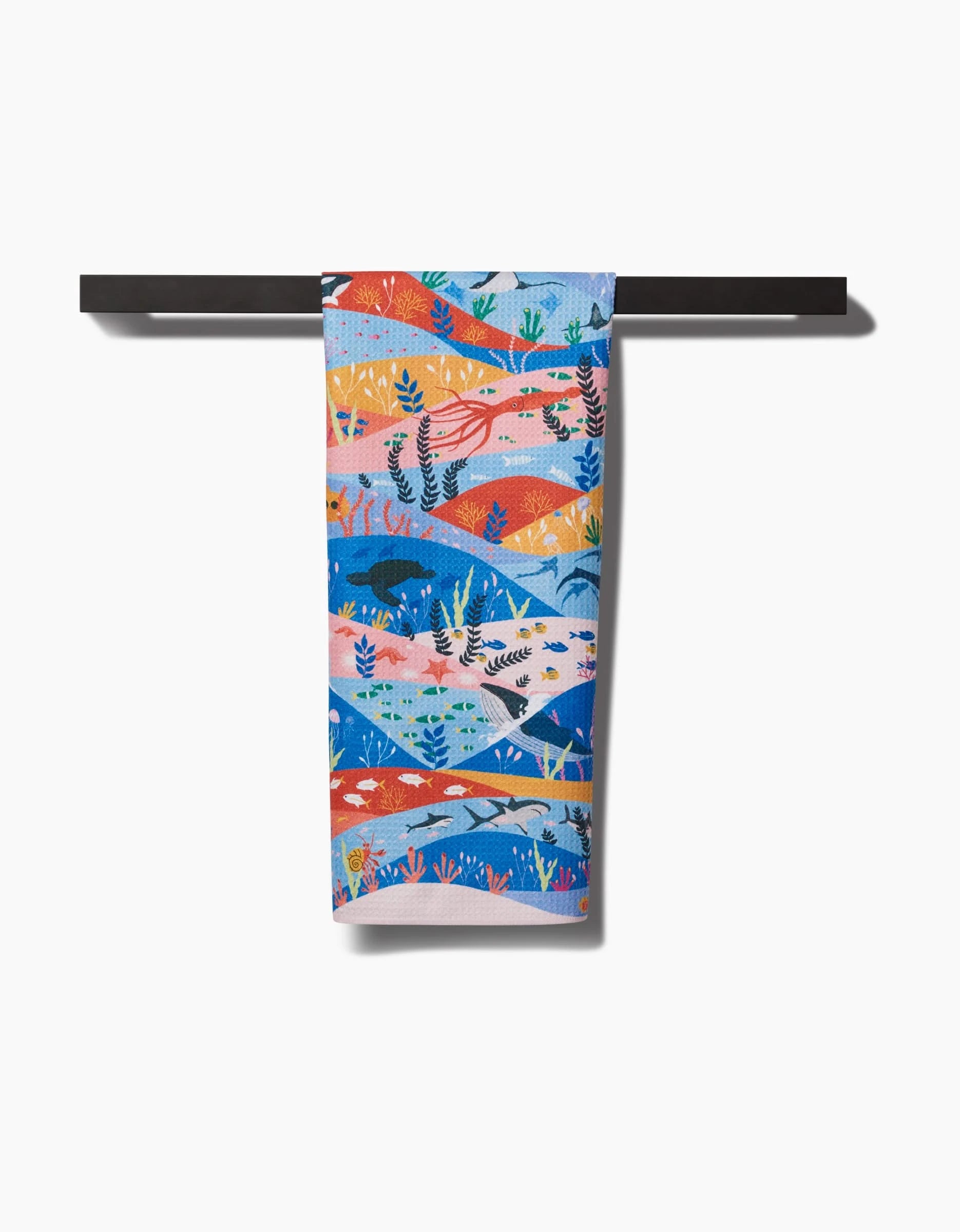 The Oceans tea towel by Ceyda Alasar, made from recycled materials, displays vibrant blue, orange, red, and pink sea life patterns and hangs over a black bar against a white background.