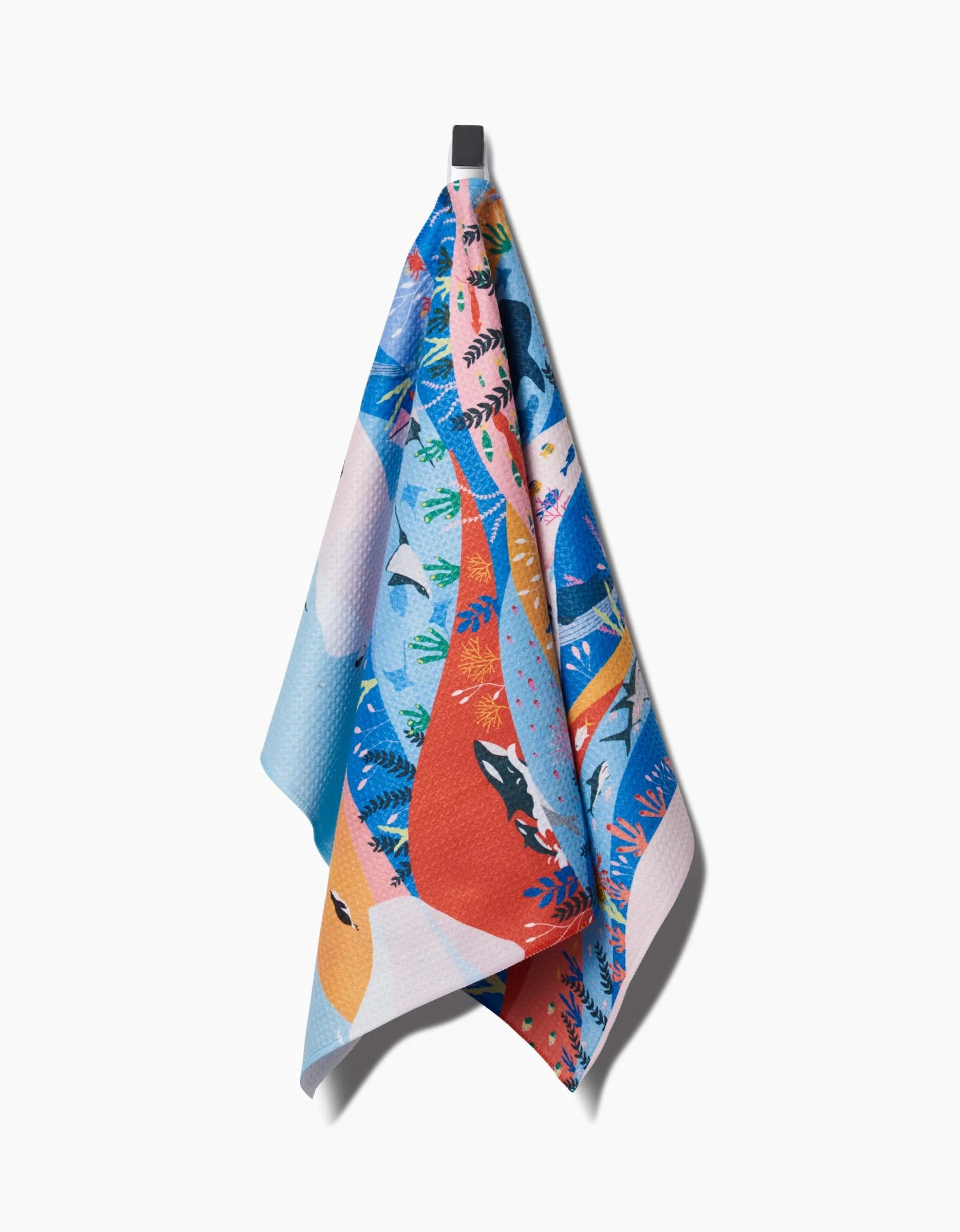 The Oceans tea towel by Ceyda Alasar features a playful animal and leaf pattern in bright oranges, blues, and pinks, crafted from Geoweave™ fabric and displayed hanging on a hook against a plain white background.