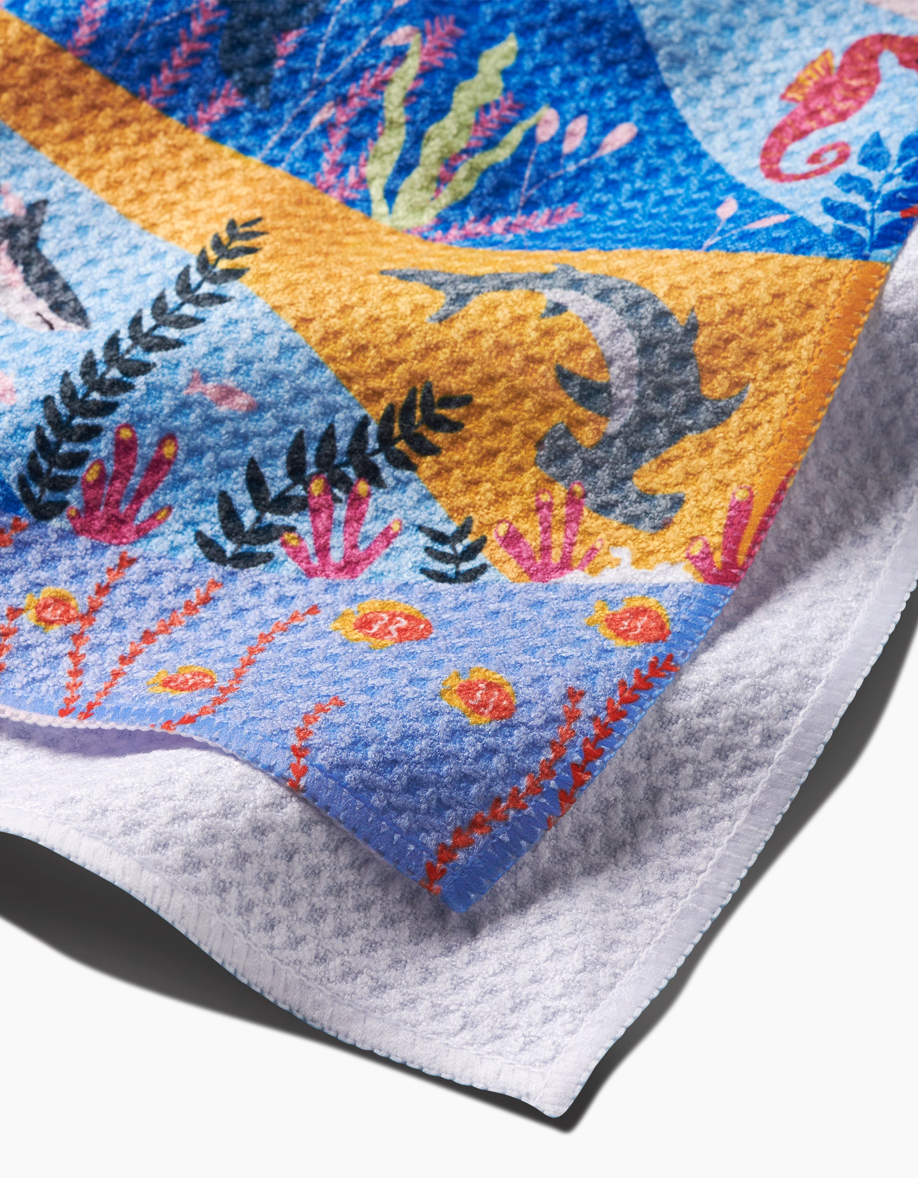 A close-up of the Oceans Tea Towel by Ceyda Alasar displays fish, seaweed, coral, seahorses, and ocean plants on textured Geoweave™ fabric, with the towel’s corner neatly folded over a white surface.