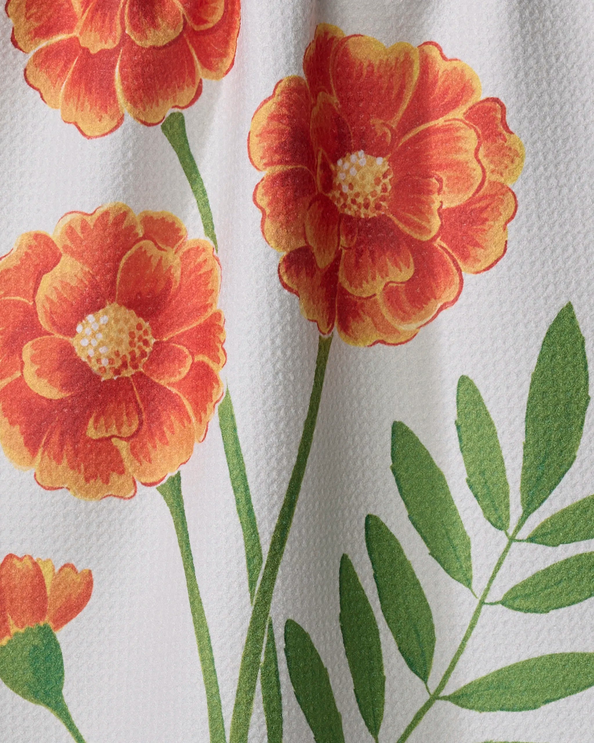 A close-up of the Linda Fox October Marigold ultra-absorbent tea towel, featuring orange and yellow marigold flowers with green leaves on a textured white background.