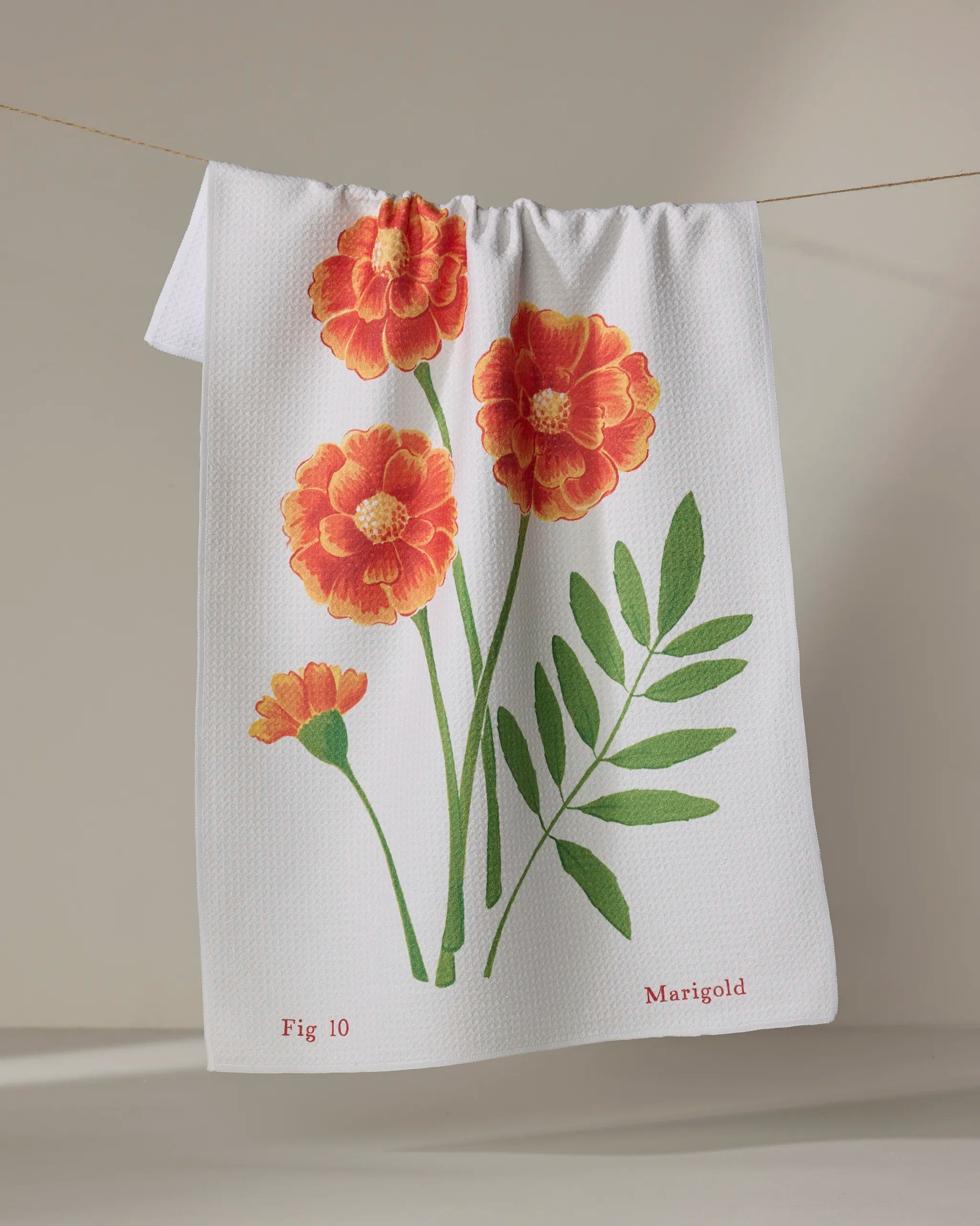 The October Marigold kitchen towel by Linda Fox, made from white recycled material, features marigold flowers with green leaves and hangs on a line. "Fig 10" and "Marigold" are printed at the bottom in red.