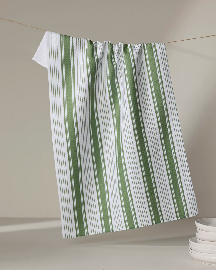 Aubrey Fairchild’s "Of the Same Stripe" tea towel, crafted from recycled materials, hangs on a clothesline above stacked white plates in the lower right corner. Its white fabric features vertical green and thin black stripes against a neutral backdrop.
