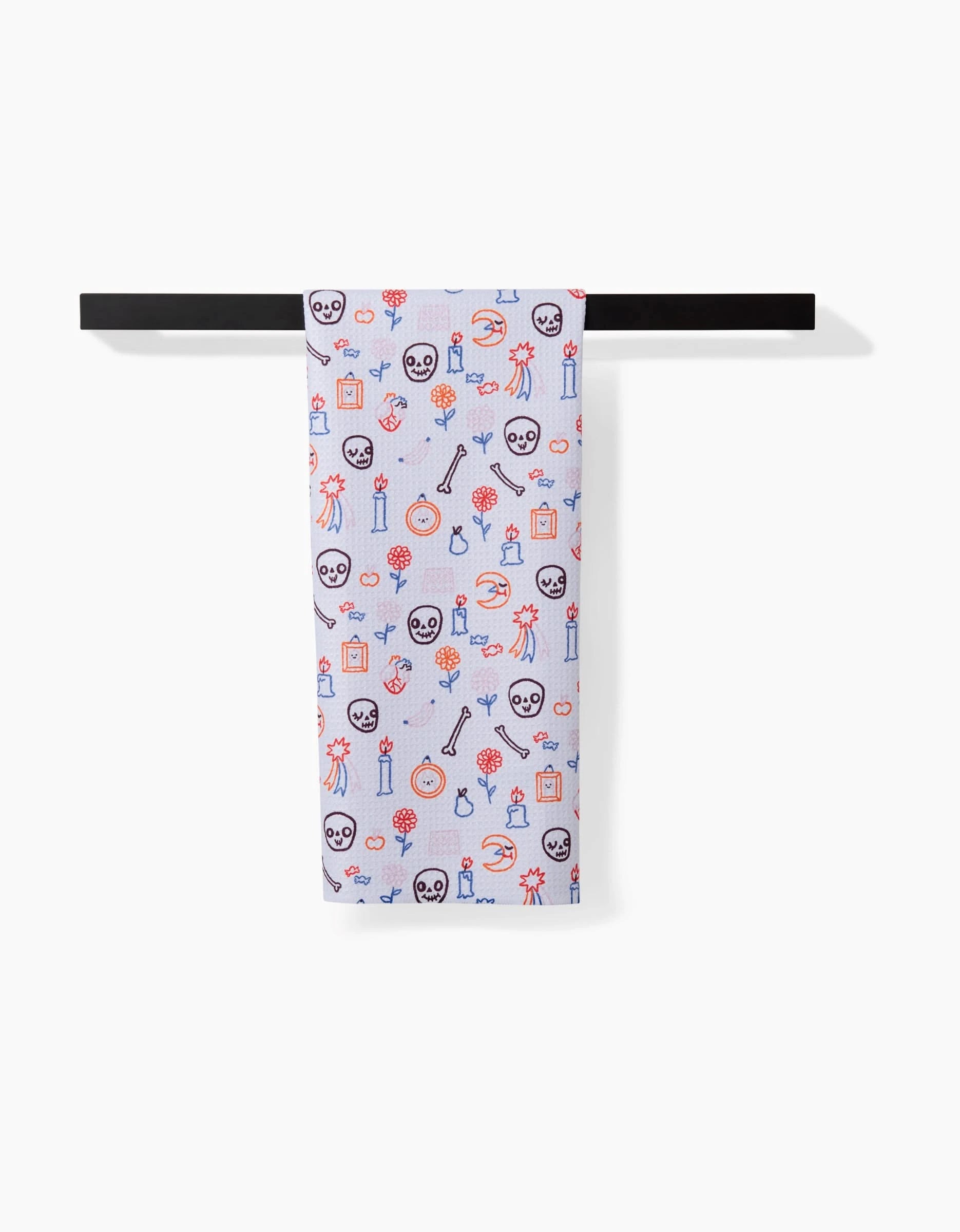 The Offerings tea towel by So Lazo, made from recycled materials, displays vibrant skulls, cacti, guitars, flowers, and candles. Shown on a black towel bar against a white backdrop.