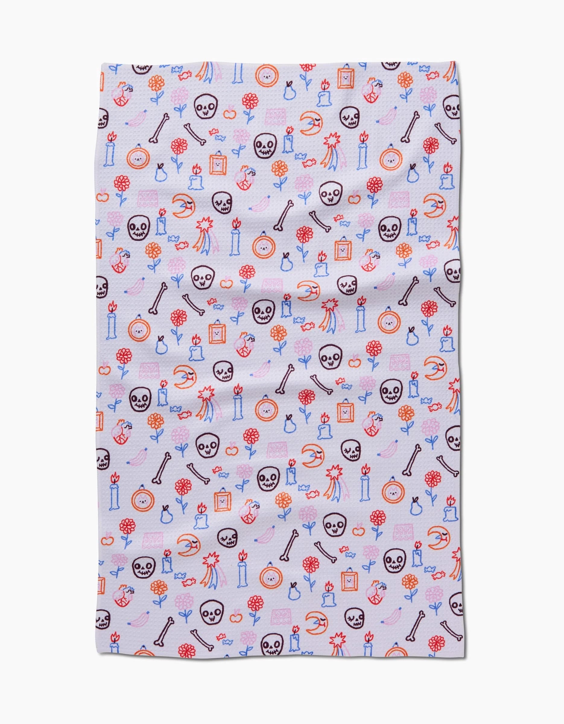The So Lazo Offerings tea towel is rectangular, made from recycled materials, and features a playful mix of cartoon bones, skulls, mushrooms, candles, potions, and flowers in vibrant reds, blues, oranges, and purples on white.