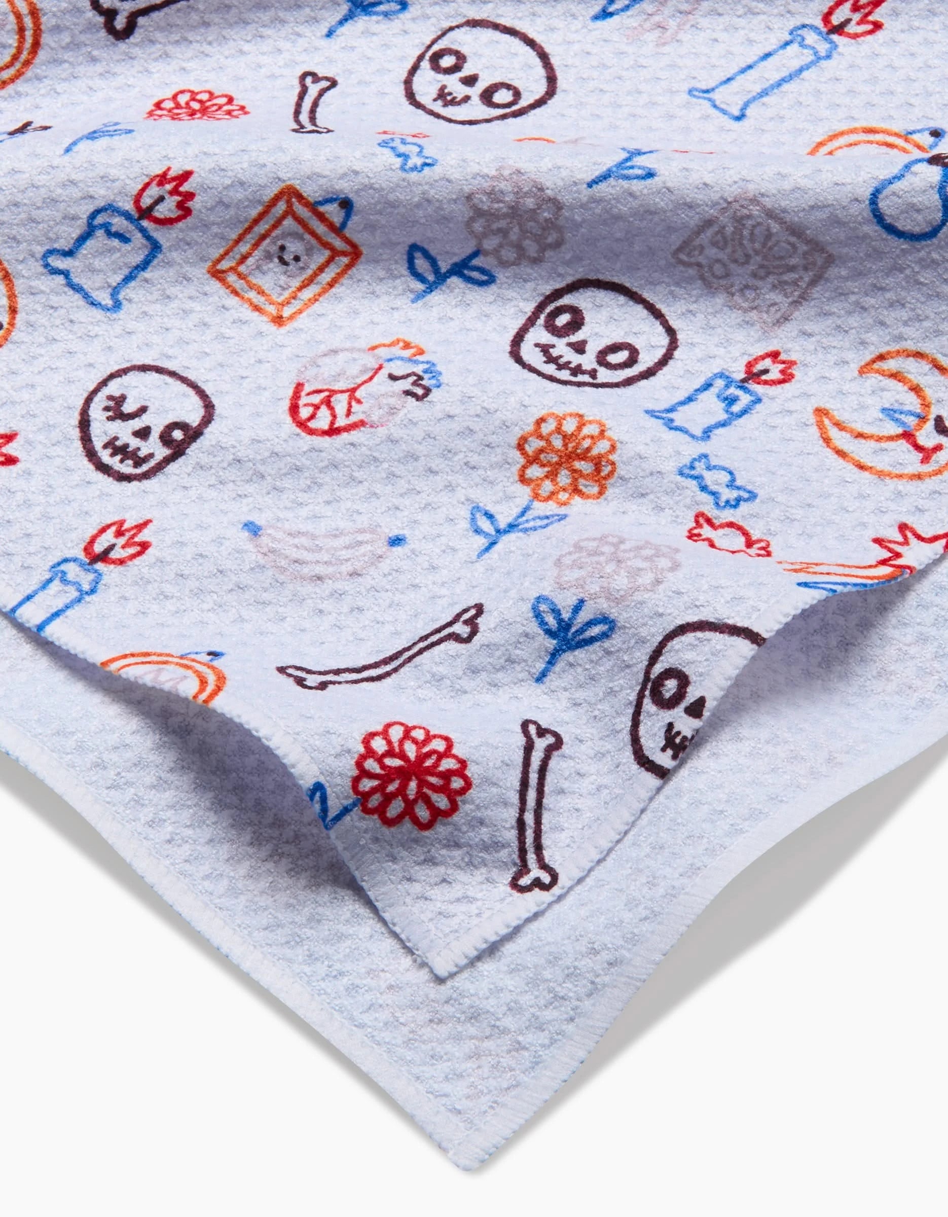 A close-up of the Offerings tea towel by So Lazo, featuring colorful skulls, bones, flowers, moons, candles, and frames on white ultra-absorbent recycled fabric; corner folded to show texture.