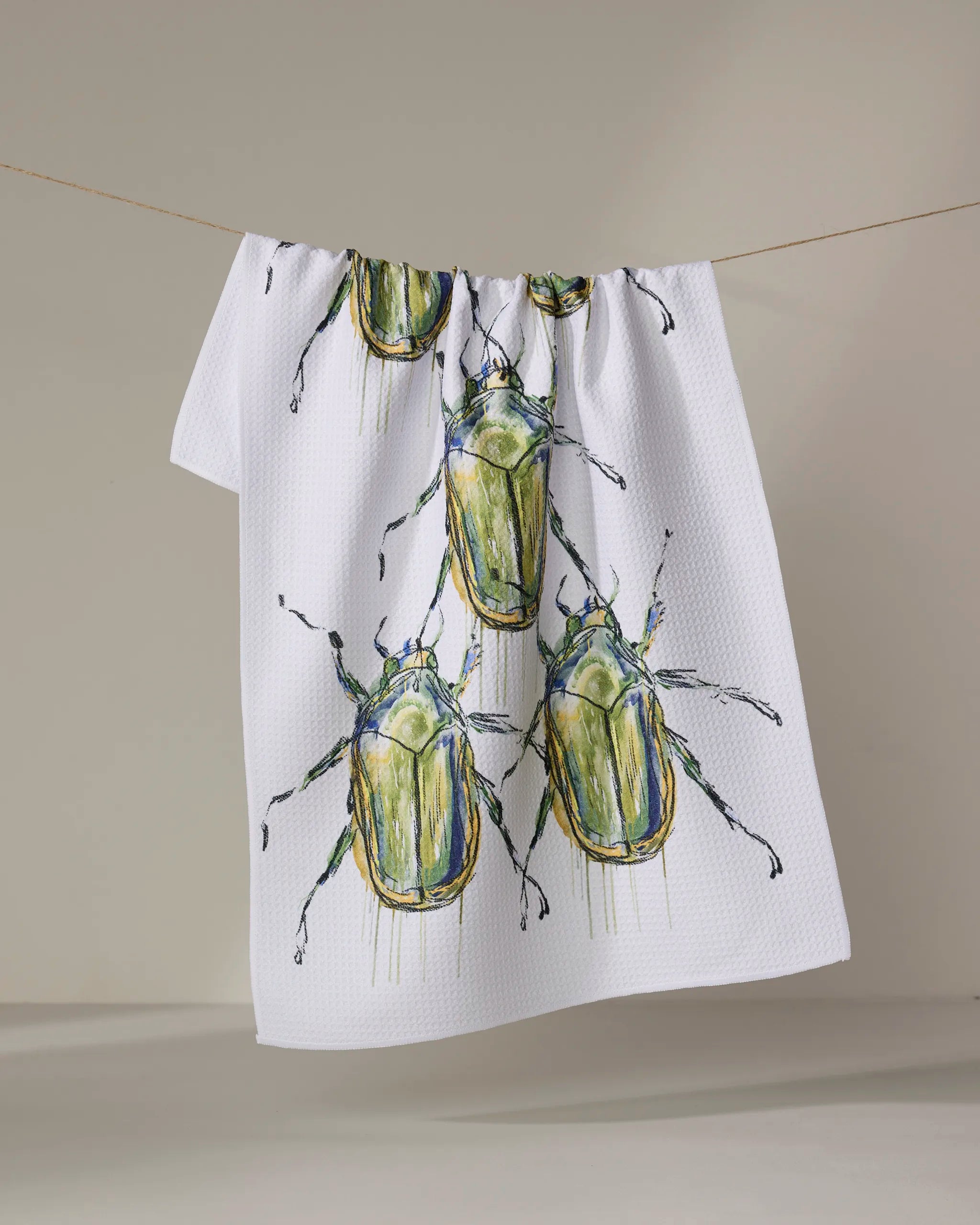 The Olive Beetles kitchen towel by Taryn Brennan features eco-friendly white fabric with large green and yellow watercolor beetles, shown hanging on a clothesline against a plain background.