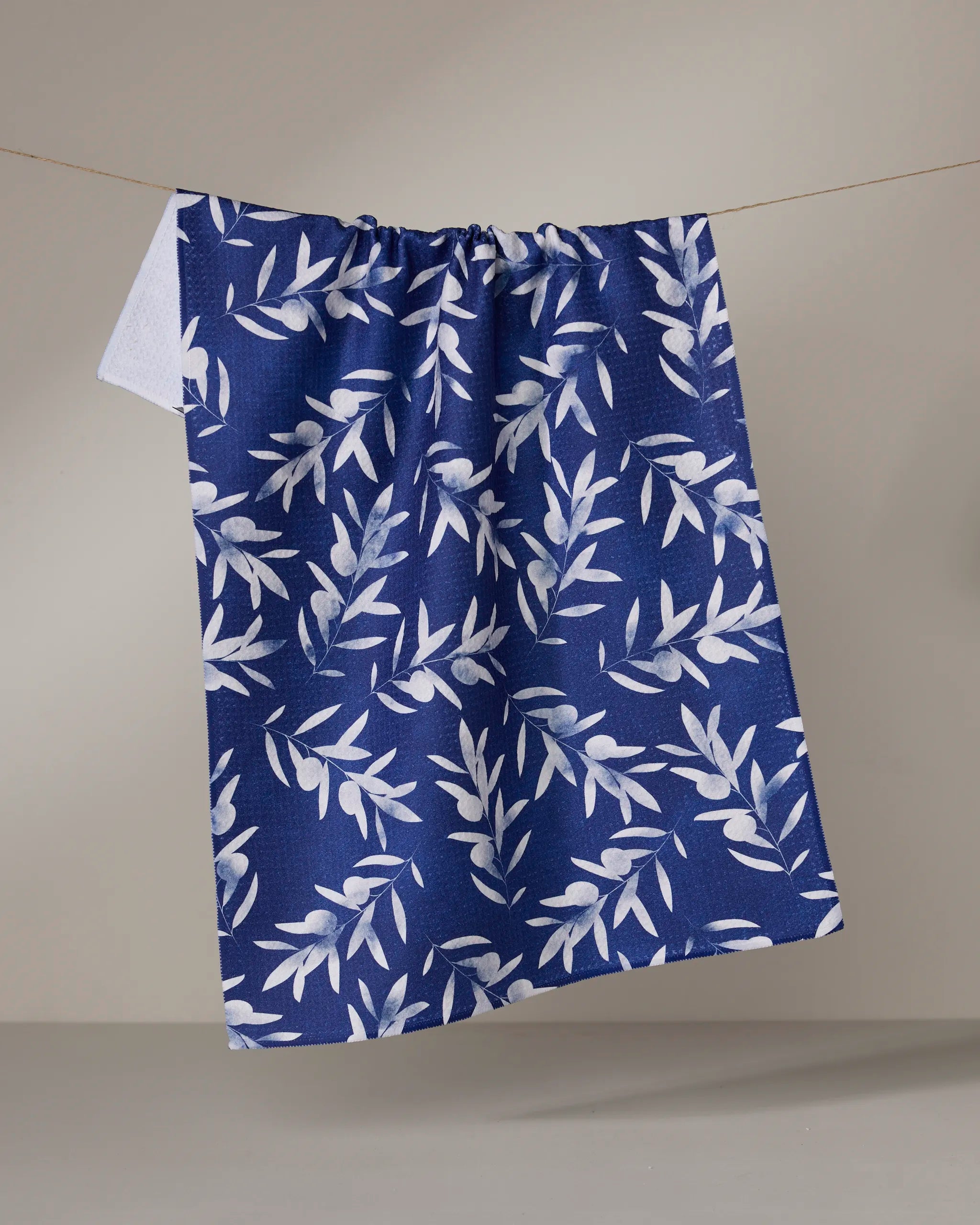 The Geometry Olive Branch kitchen towel, featuring a white leafy pattern on blue and made from recycled materials, hangs on a clothesline against a light background.
