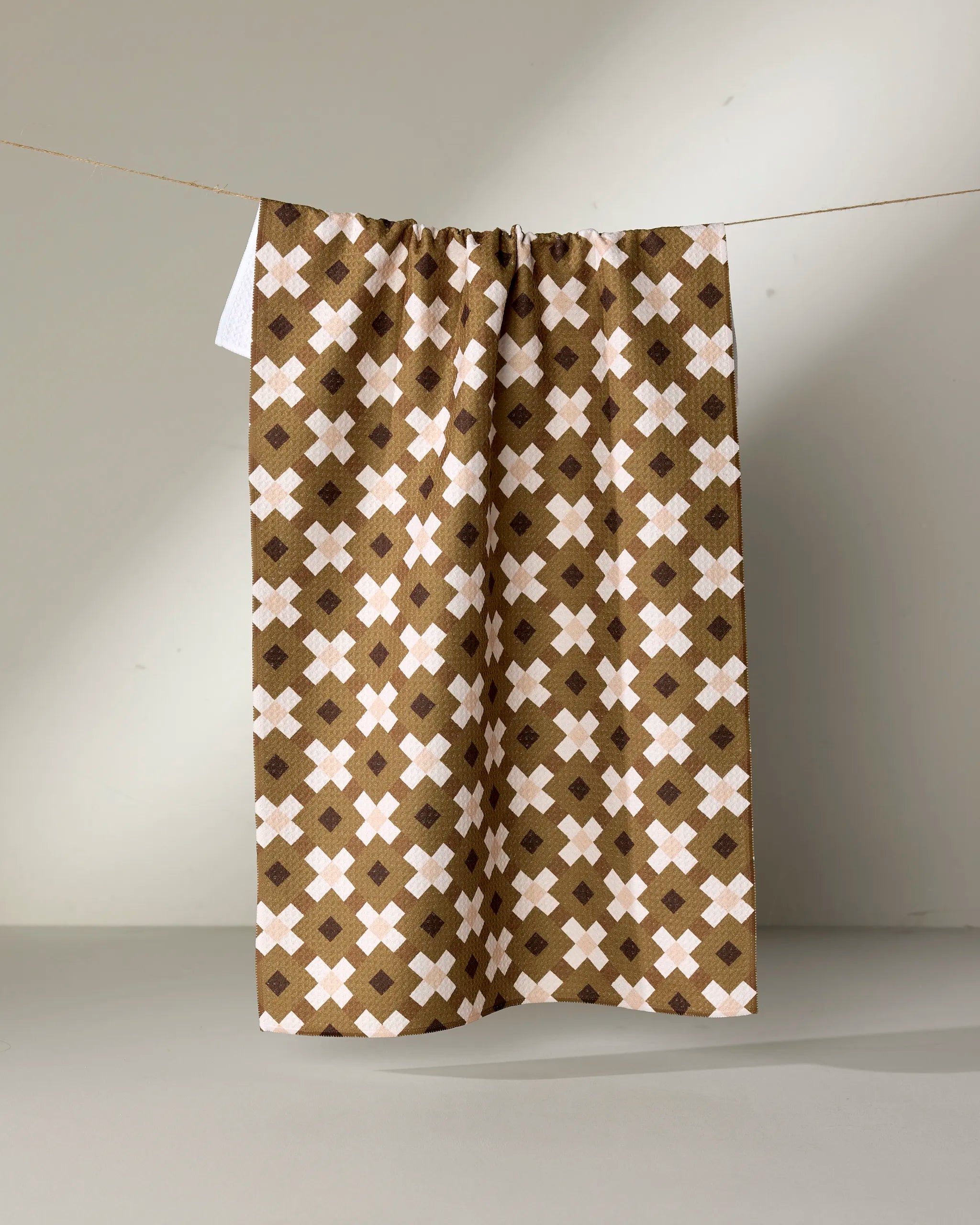 The Olive Geo recycled kitchen towel by Teri the Butter, featuring a brown and white geometric cross pattern, hangs on a string against a neutral background.
