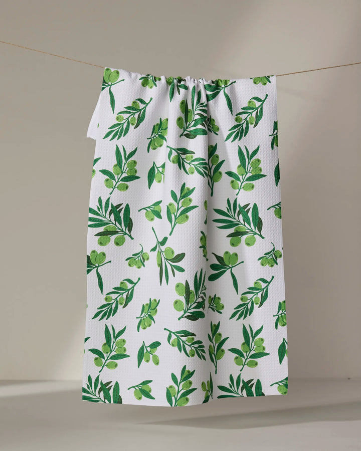 The Olive Grove tea towel by Vaishali Design Studio, showcasing a green leafy and olive branch pattern, hangs on a thin clothesline against a simple, light background.