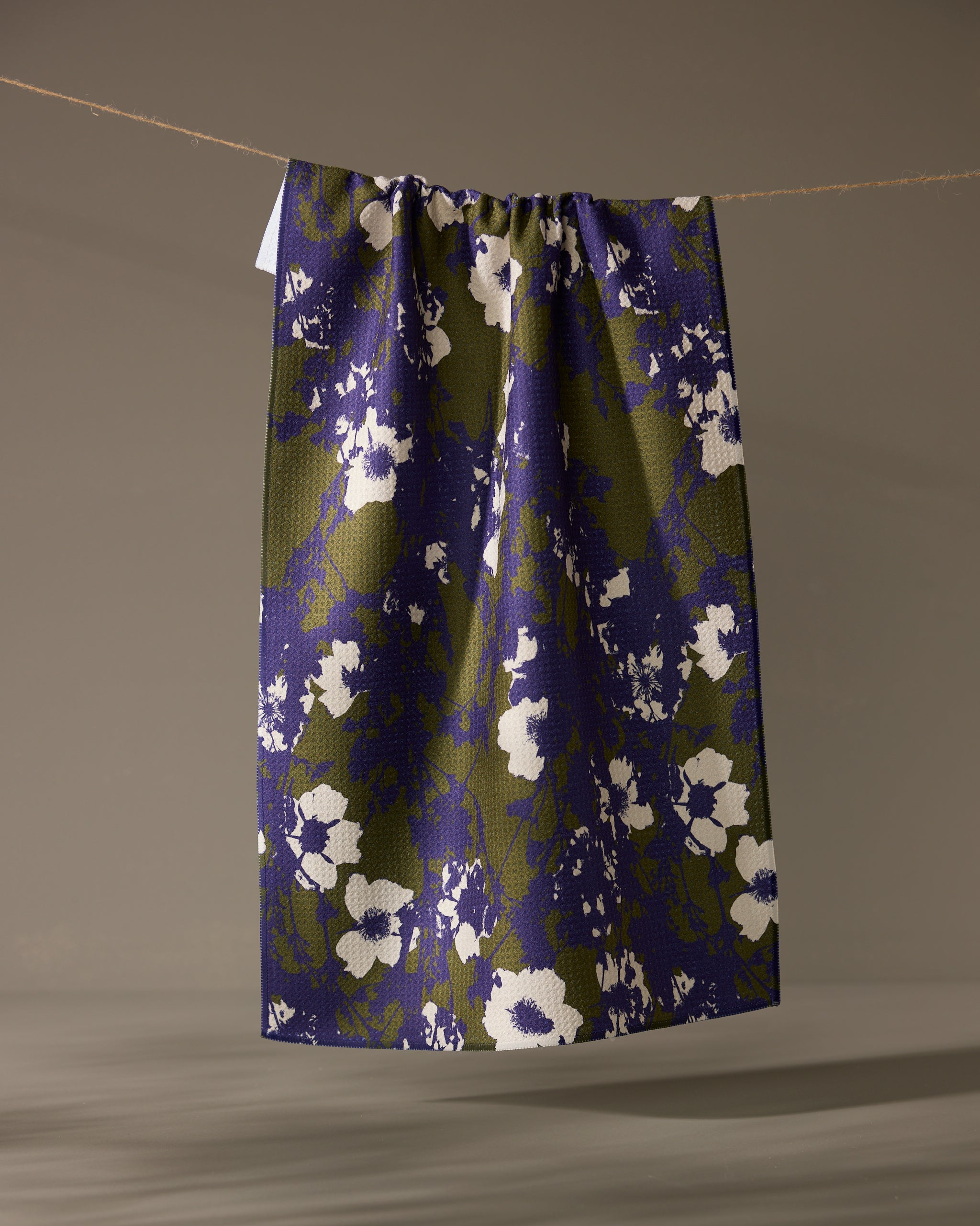 The Oliveberry skirt by Geometry, knee-length with a gathered waistband, made from quick-drying recycled materials and adorned with a blue and olive green abstract floral print, hangs from a clothesline against a neutral background.