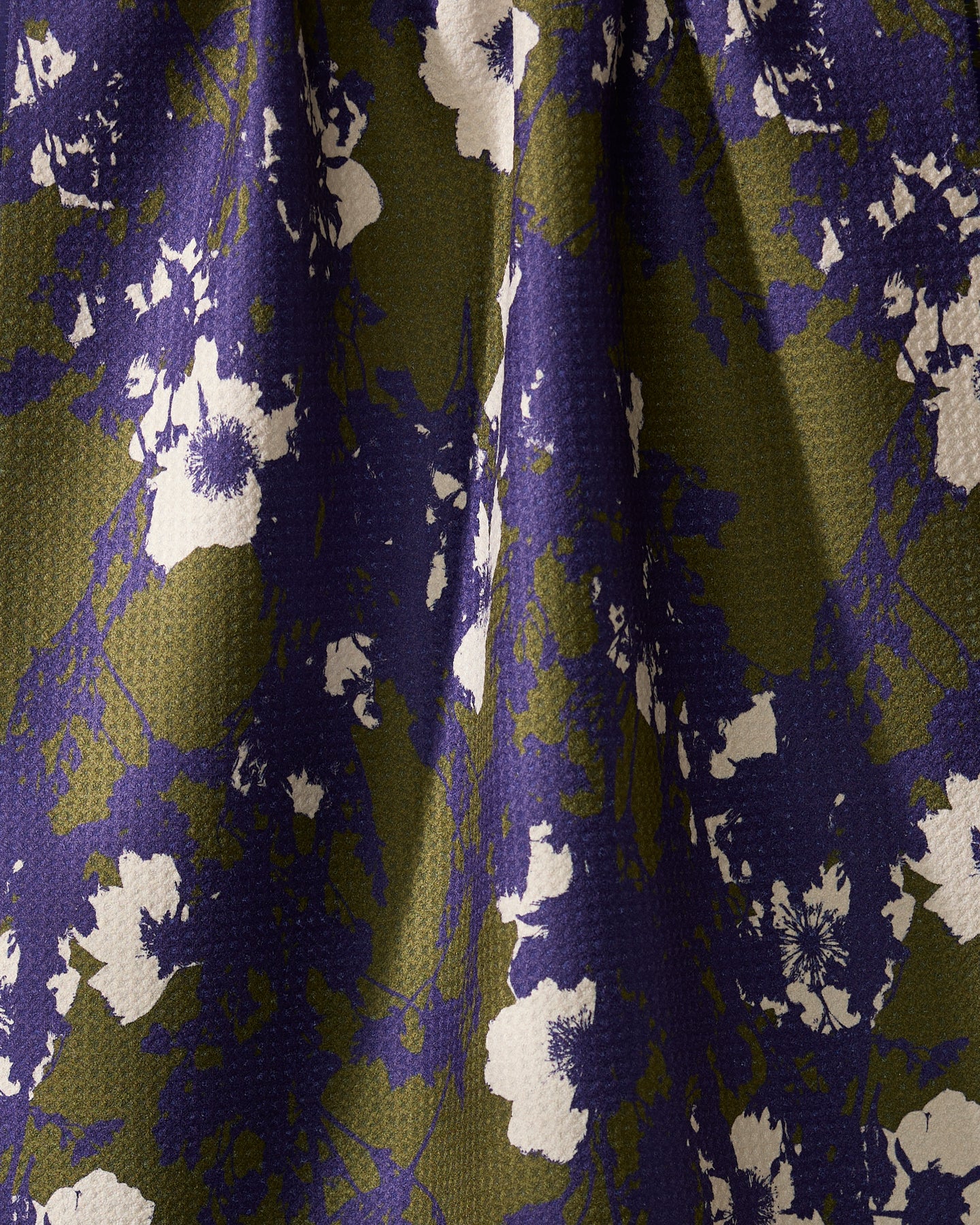 A close-up of Geometrys Oliveberry tea towel, crafted from quick-drying recycled materials. It features a softly pleated, textured surface with an abstract floral pattern in white, purple, and olive green.