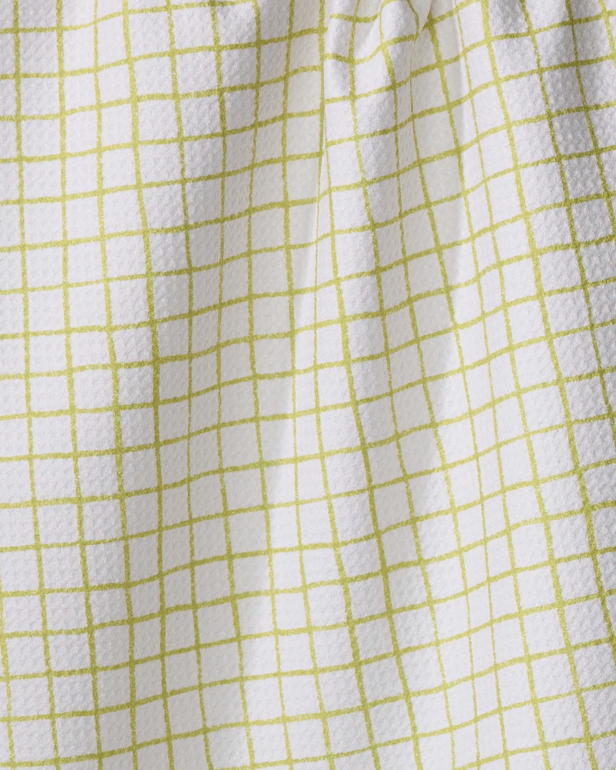 The On the Lawn kitchen towel by Kayla Ann is white with a textured finish and a grid pattern of thin light green lines, creating soft folds and subtle shadows.